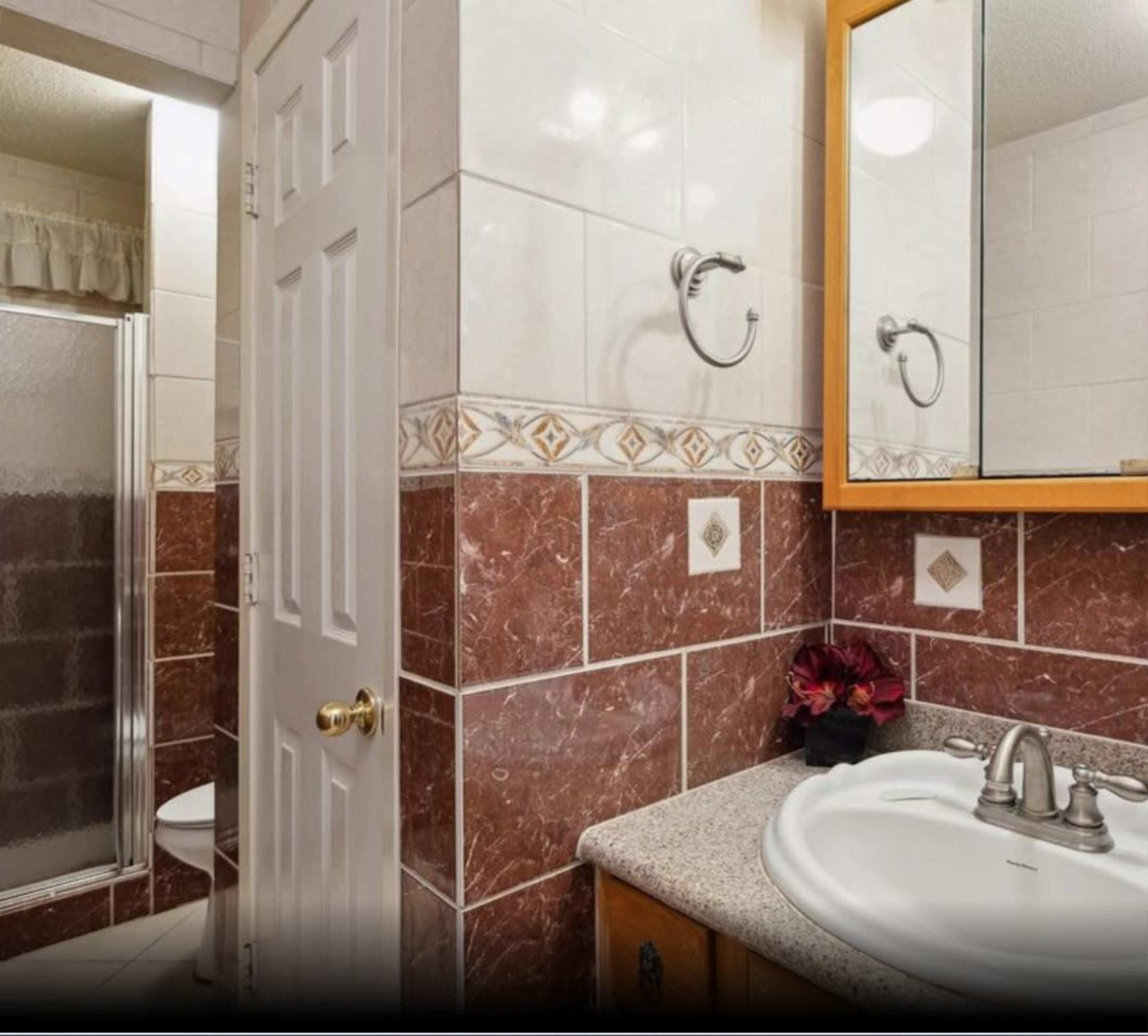 The image shows a bathroom featuring a tiled wall, a shower stall, a sink with a mirror above, and a decorative vase.