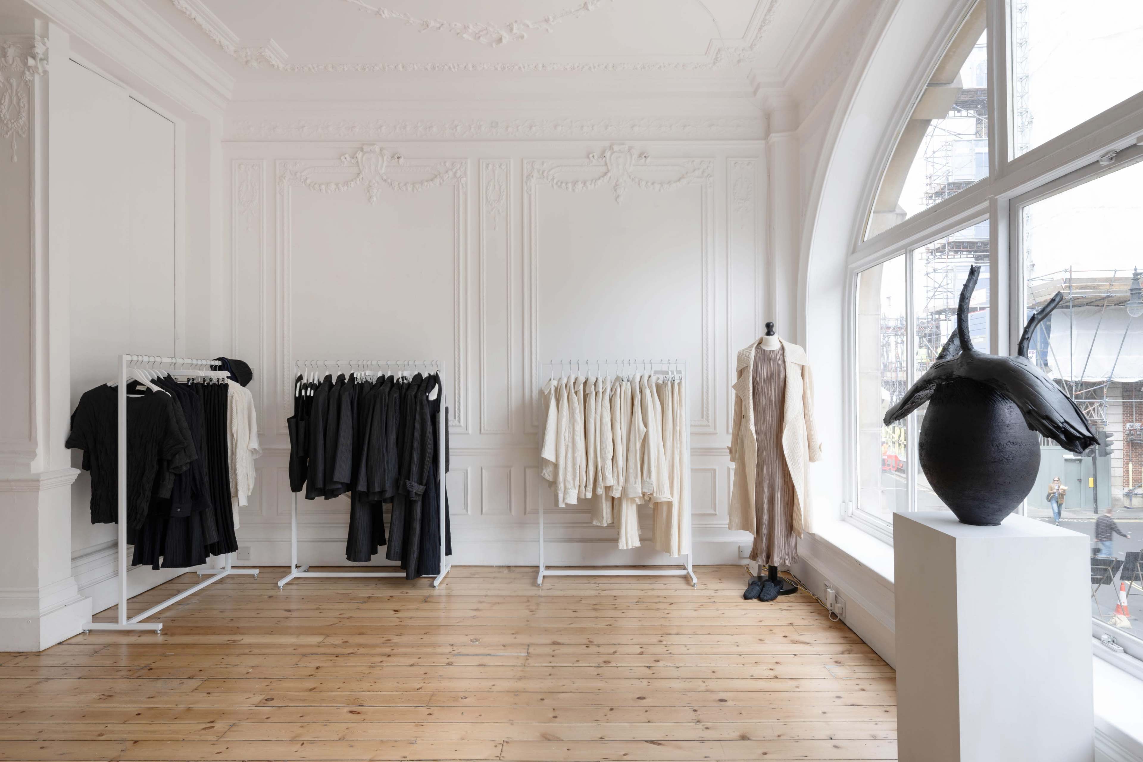 The image shows a minimalist clothing store interior with racks of black and white garments against white walls and wooden flooring, complemented by a decorative sculpture on a pedestal.