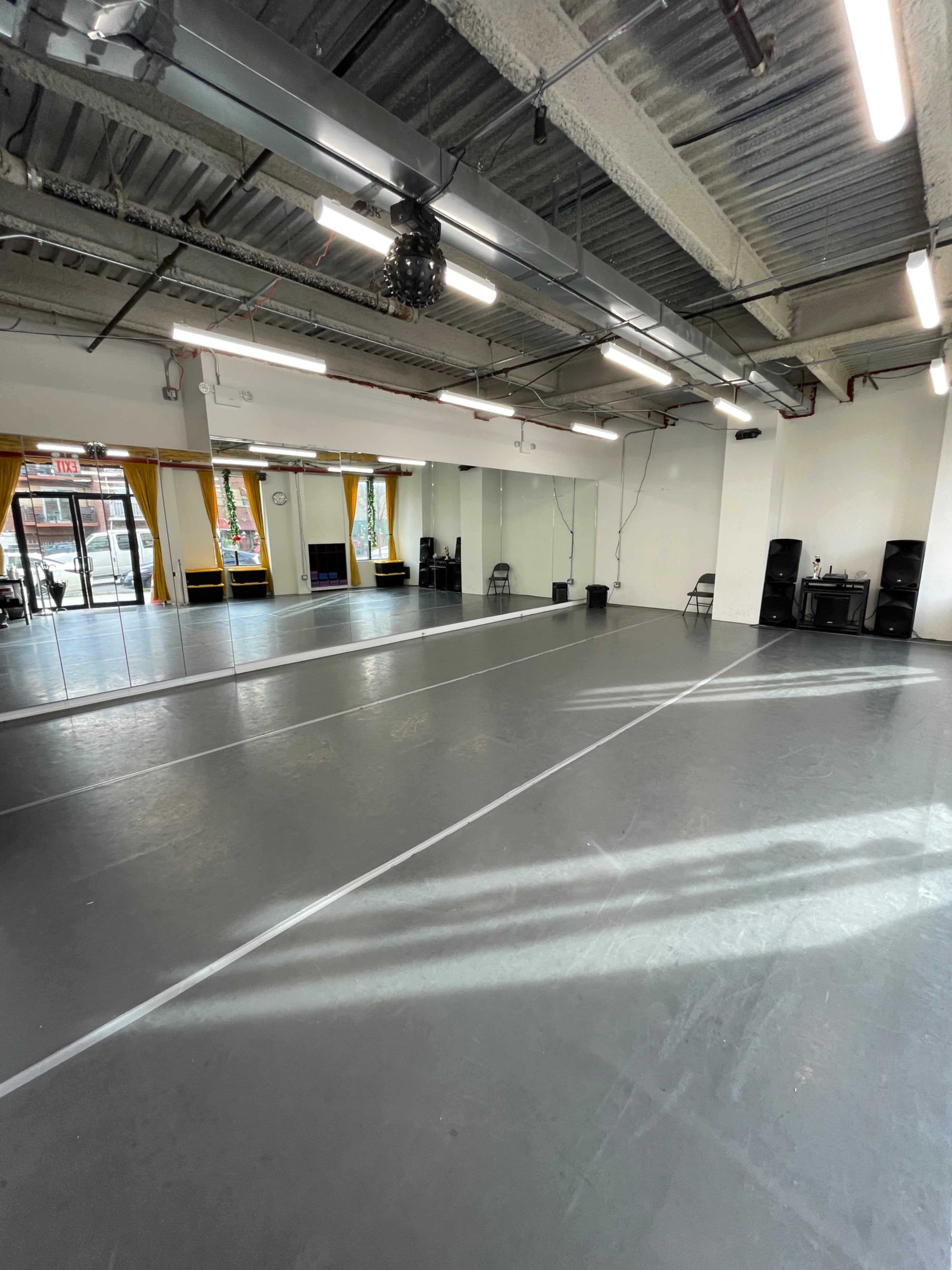 A spacious dance studio with gray flooring, wall mirrors, and large windows allowing natural light to enter.