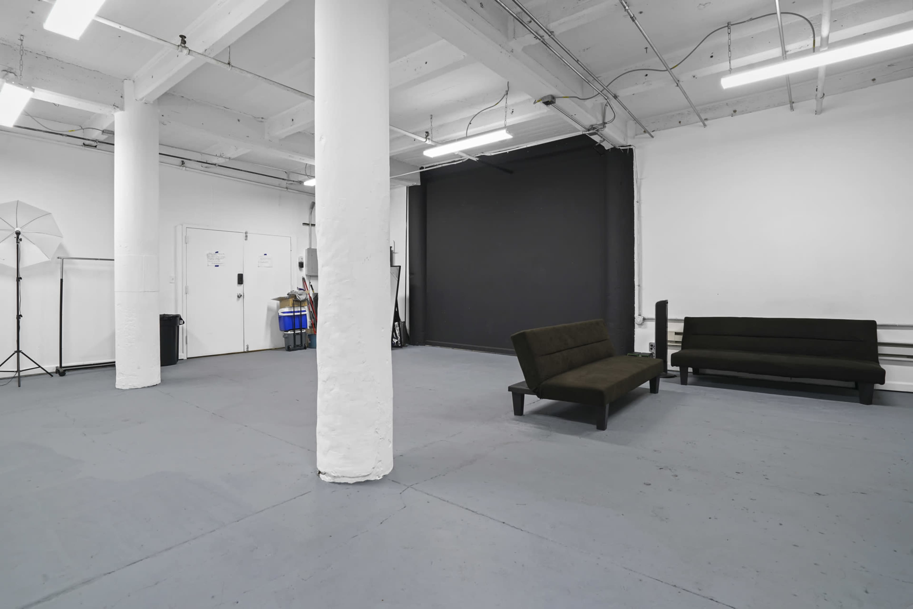 A spacious, empty room with gray flooring, white pillars, a black wall, and two dark couches positioned against one side.