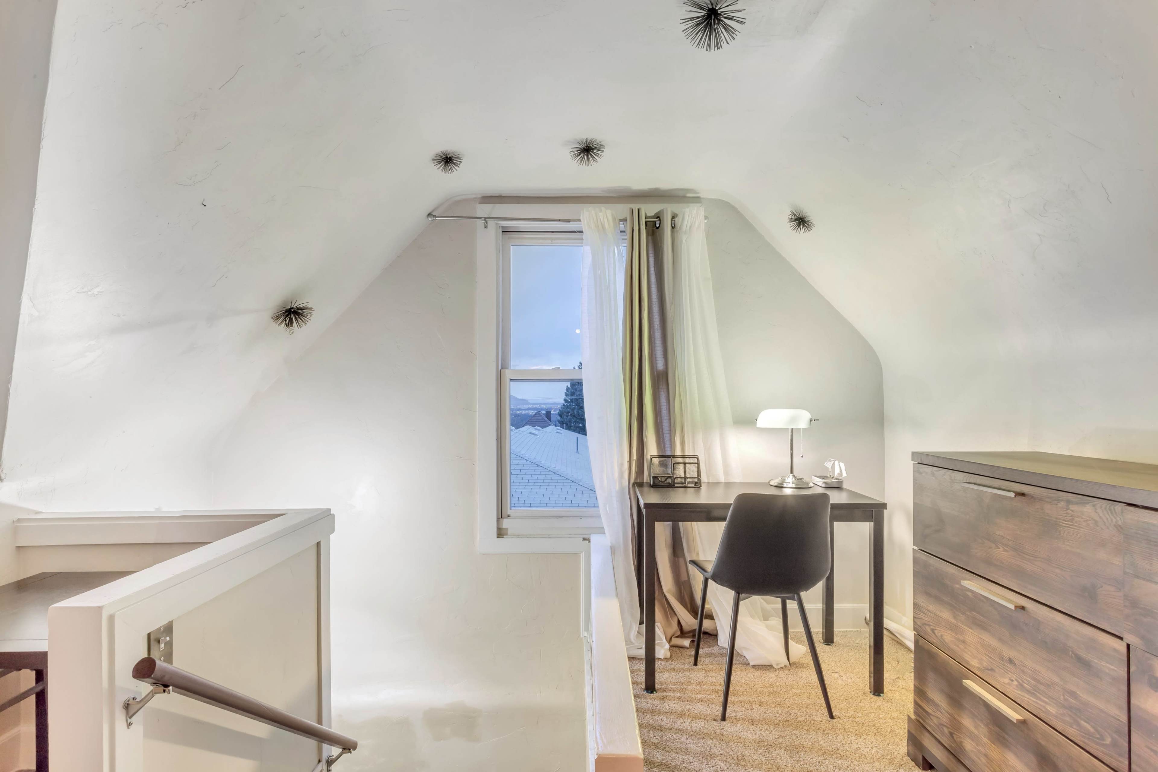An attic room with a desk and chair beside a window, featuring a wooden dresser and a textured carpeted floor.
