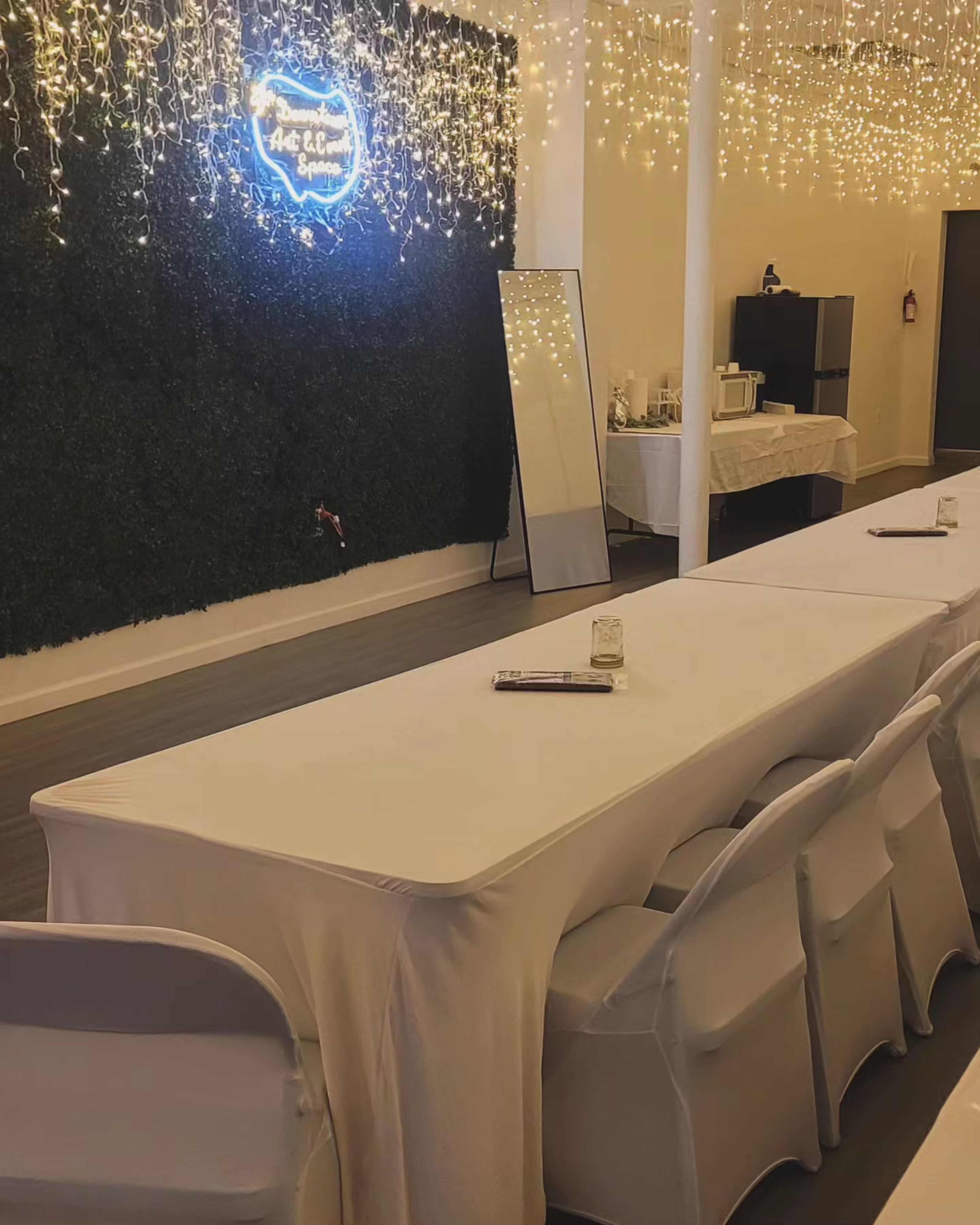 A decorated event space with a long table covered in a white tablecloth, surrounded by white chairs, and a wall adorned with greenery and string lights.