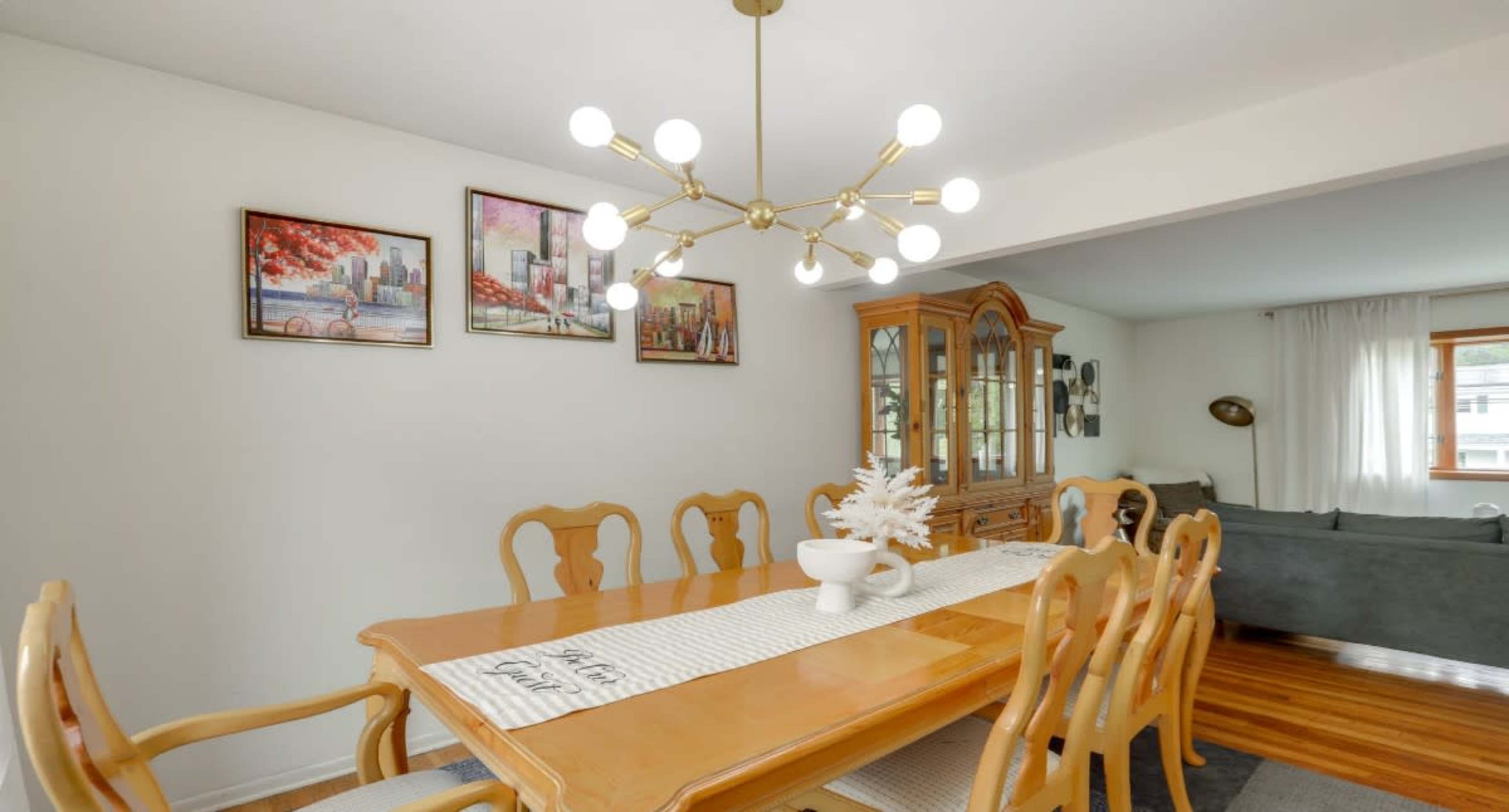The image shows a well-lit dining room featuring a wooden dining table surrounded by chairs, adorned with a decorative table runner and an artistic centerpiece, alongside a china cabinet and colorful wall art.
