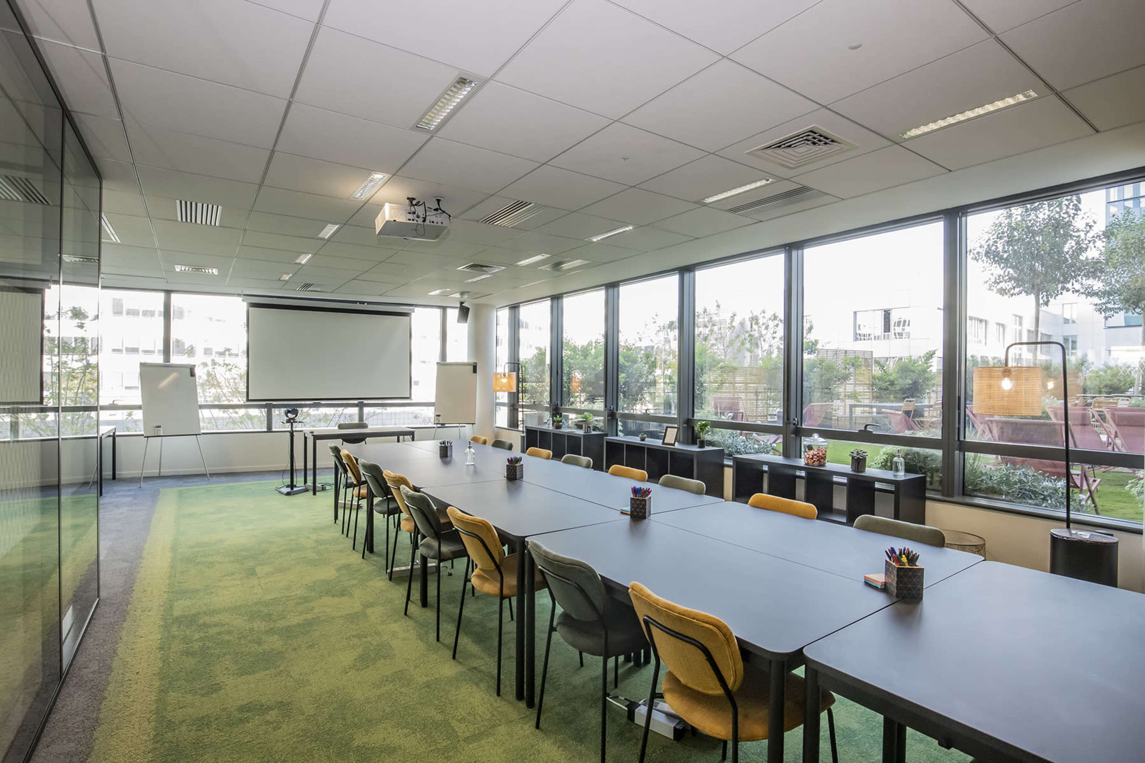 A spacious meeting room with a long table, chairs, and large windows overlooking a garden area.