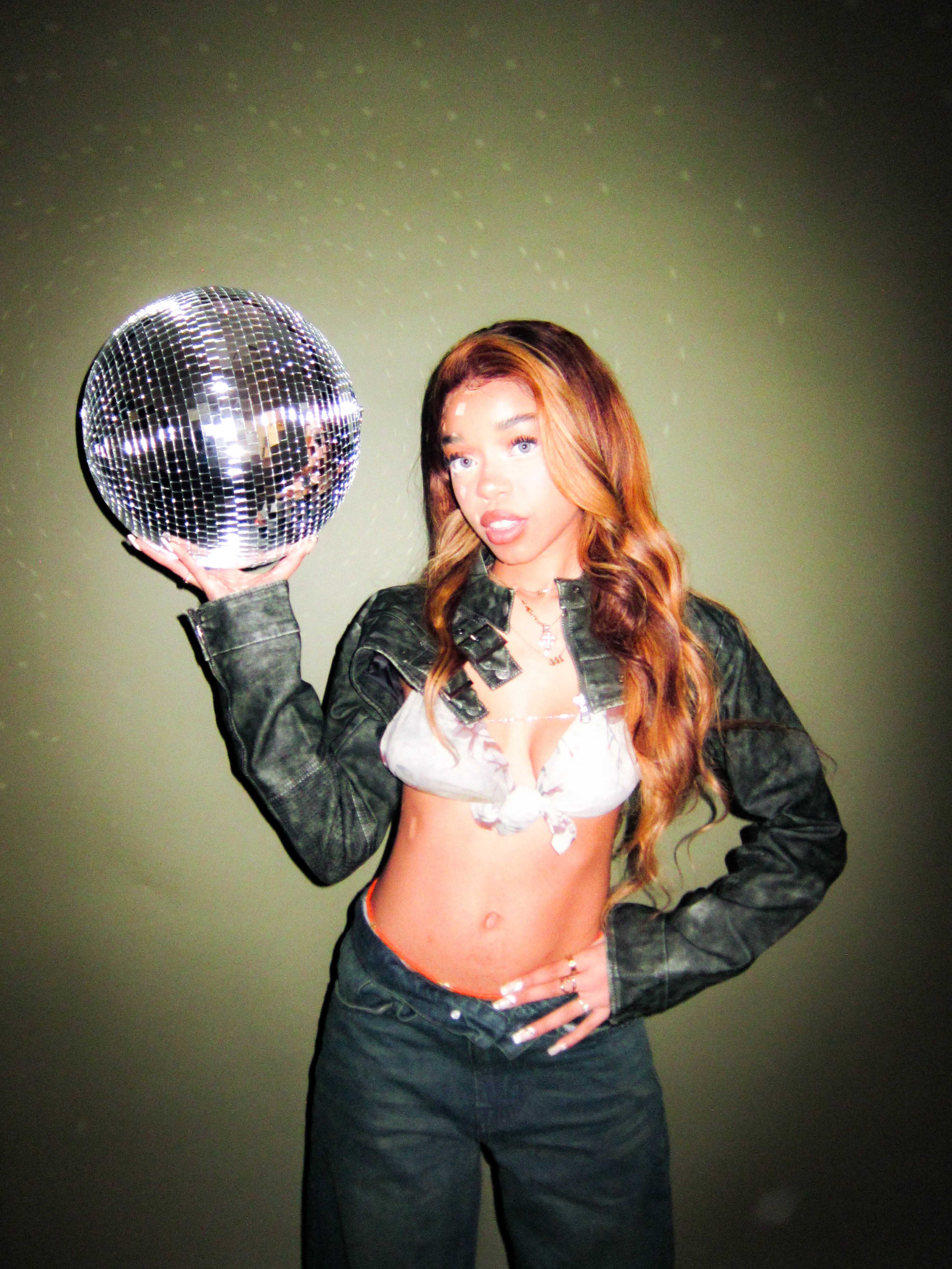 A person holds a disco ball while wearing a cropped jacket and pants against a plain green background.
