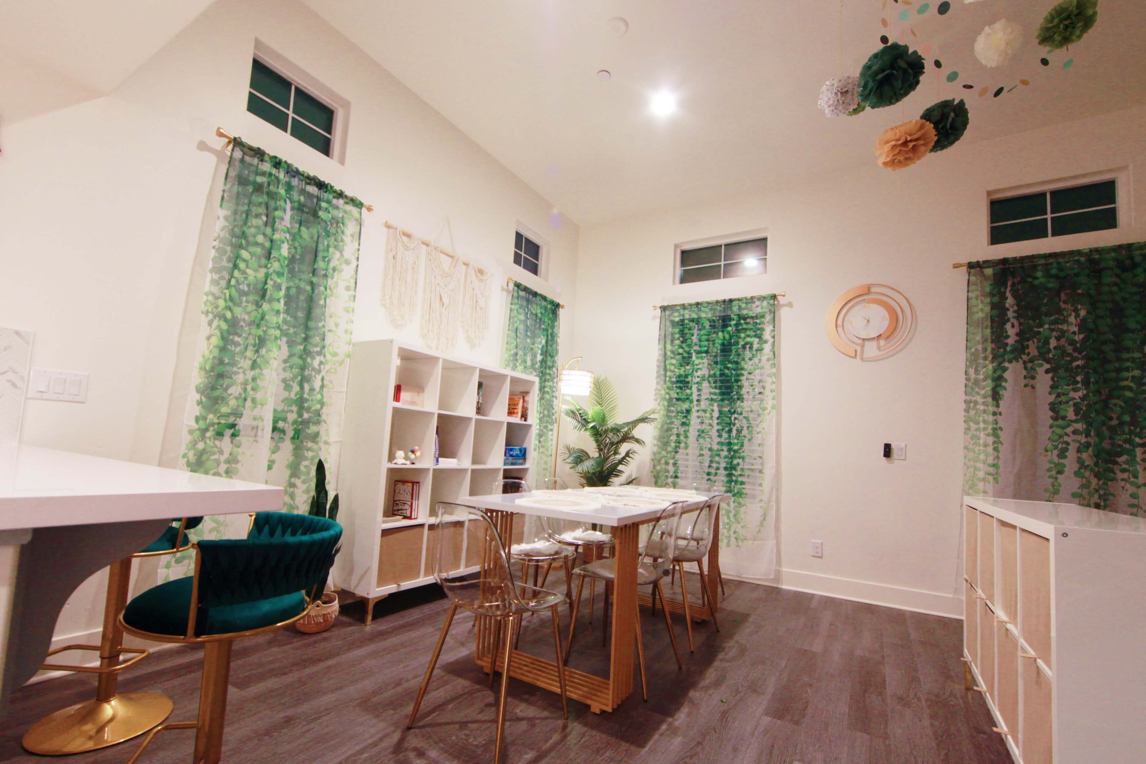 The image shows a well-lit room featuring a dining area with a white table and clear chairs, surrounded by green decorative curtains and plants.