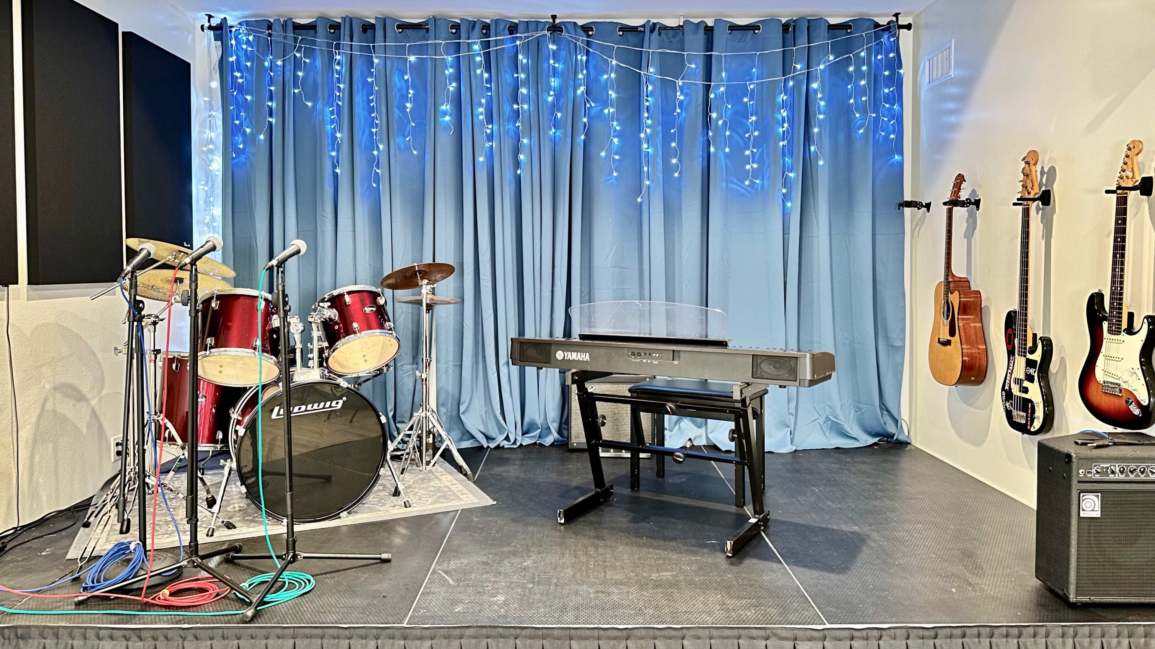 A music stage setup with a drum kit, a keyboard on a stand, and several guitars mounted on a wall, all against a backdrop of blue curtains and fairy lights.