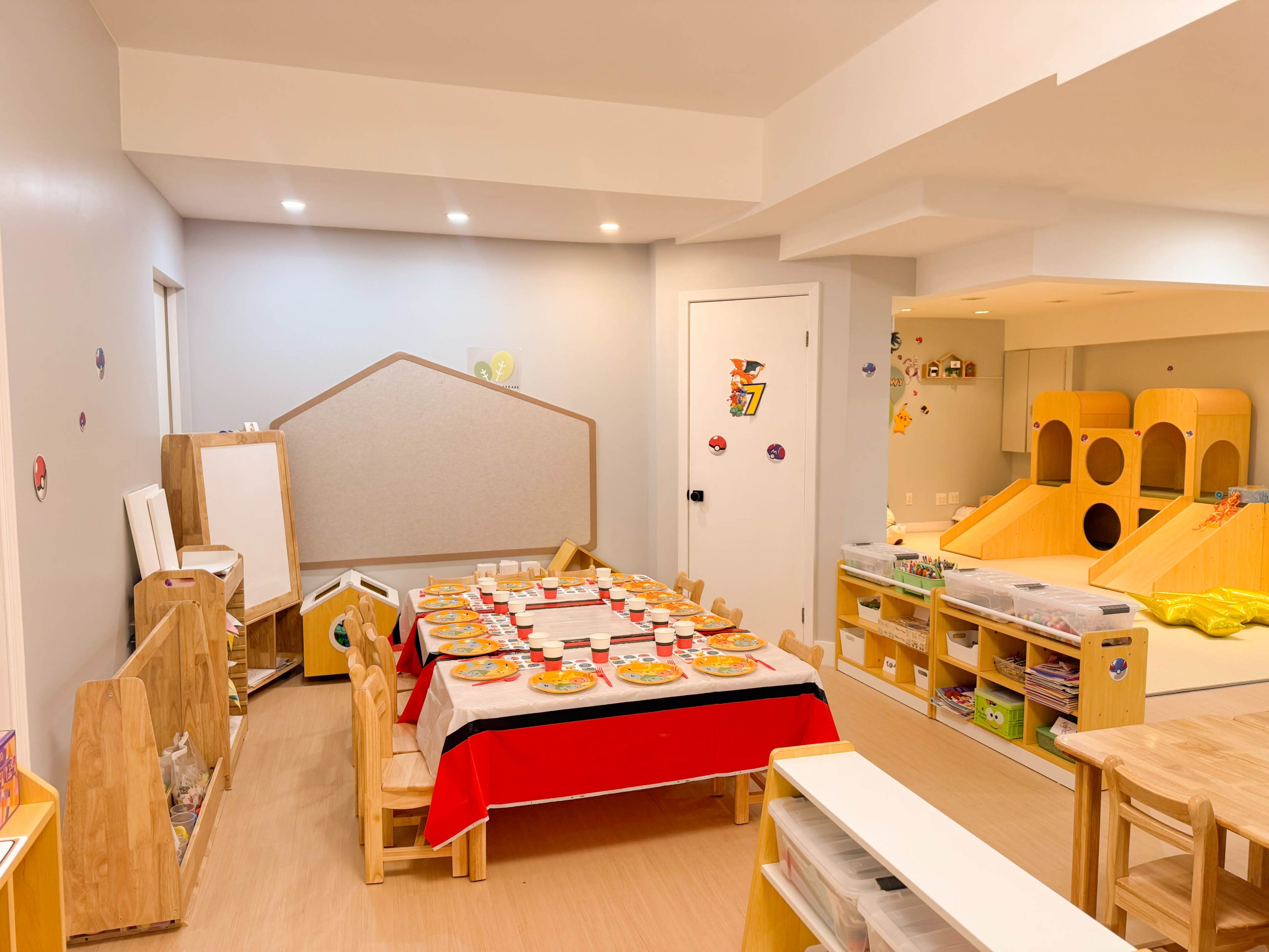 A brightly lit classroom features a red table set with colorful plates, a whiteboard, and play areas with toys and slides.