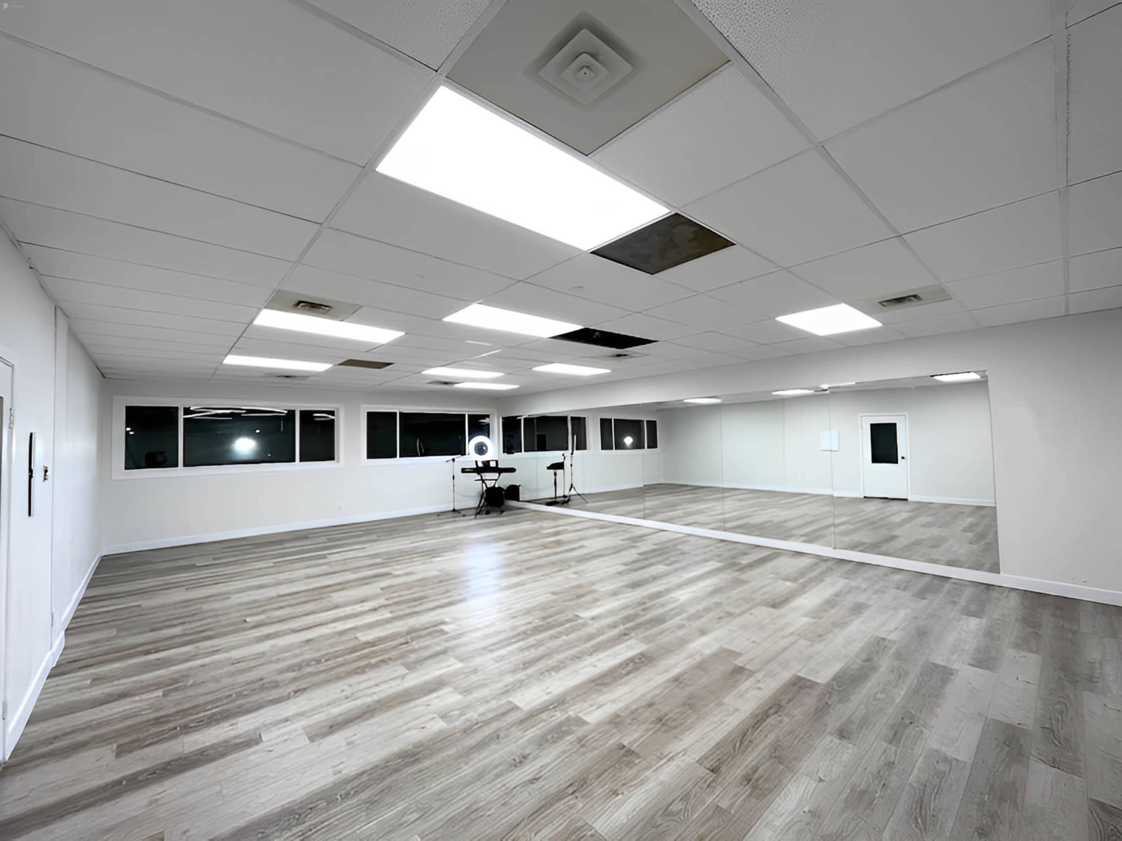 A spacious, well-lit room with wooden flooring, large mirrors, and fluorescent ceiling lights.