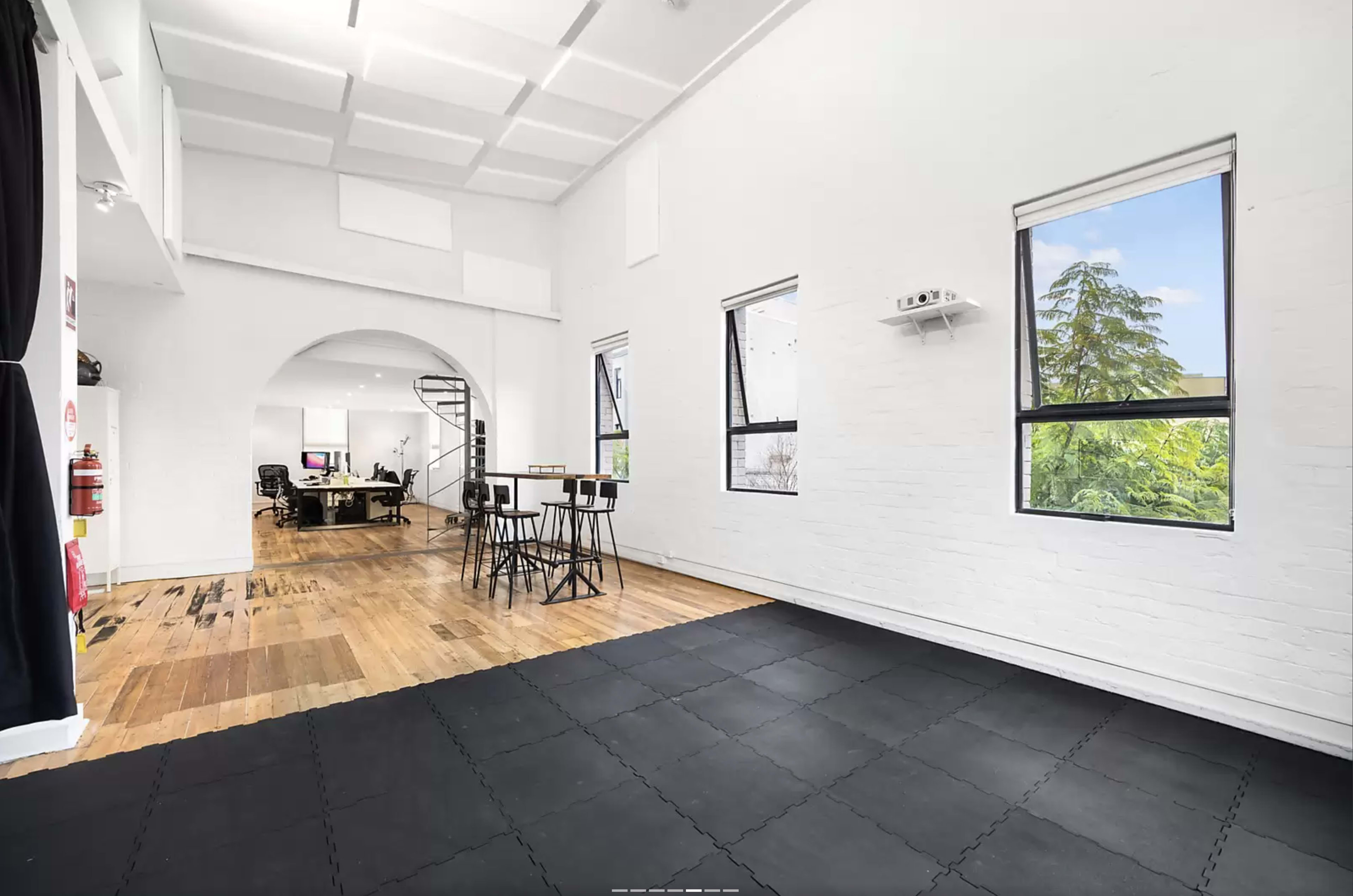 The image shows a spacious, bright room with wooden flooring, large windows, and a black rubber mat area, along with a staircase leading to an upper level and a workspace in the background.