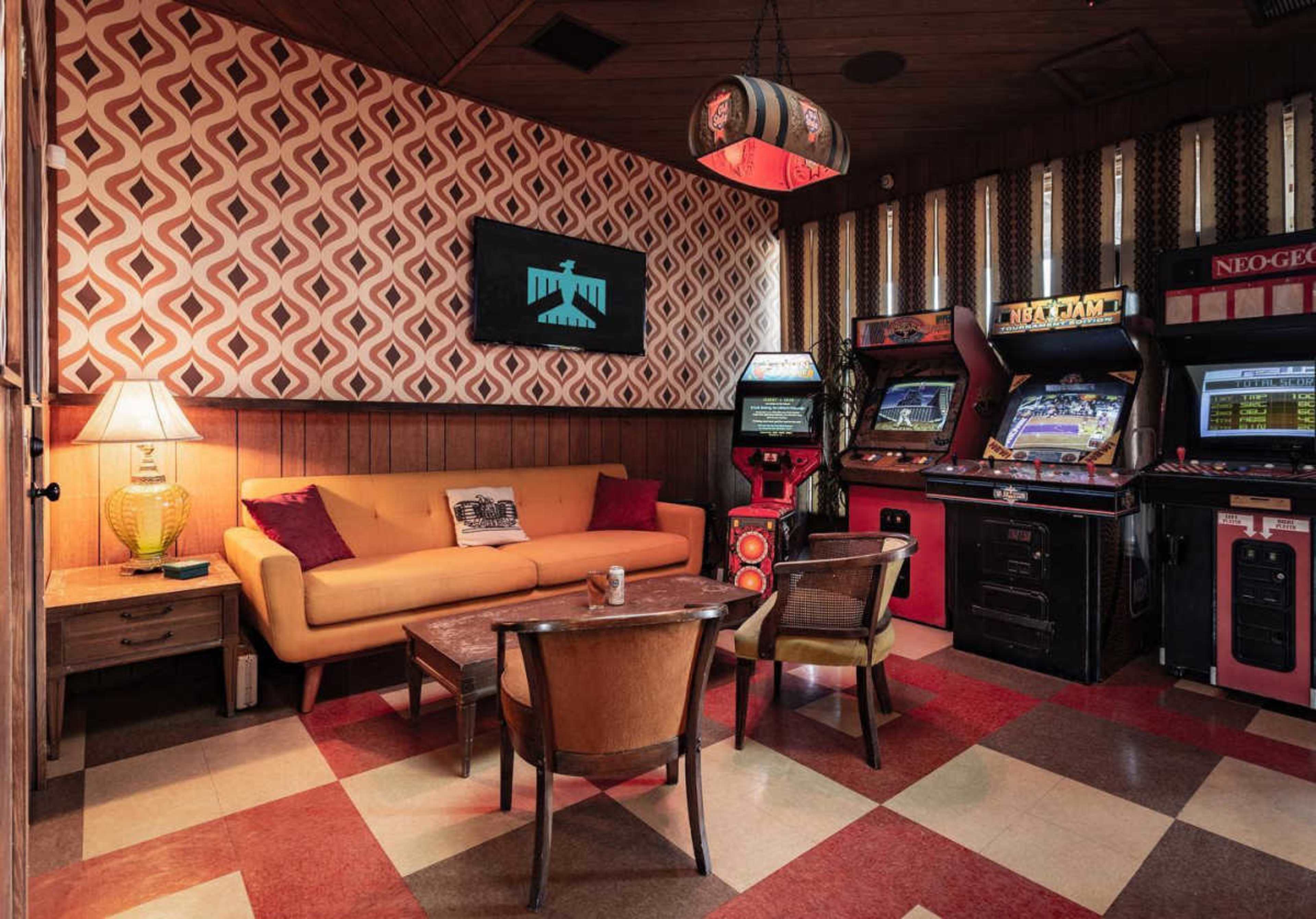 The image shows a retro-style game room featuring a brown sofa, two chairs, a wooden table, and several vintage arcade machines against a patterned wall.