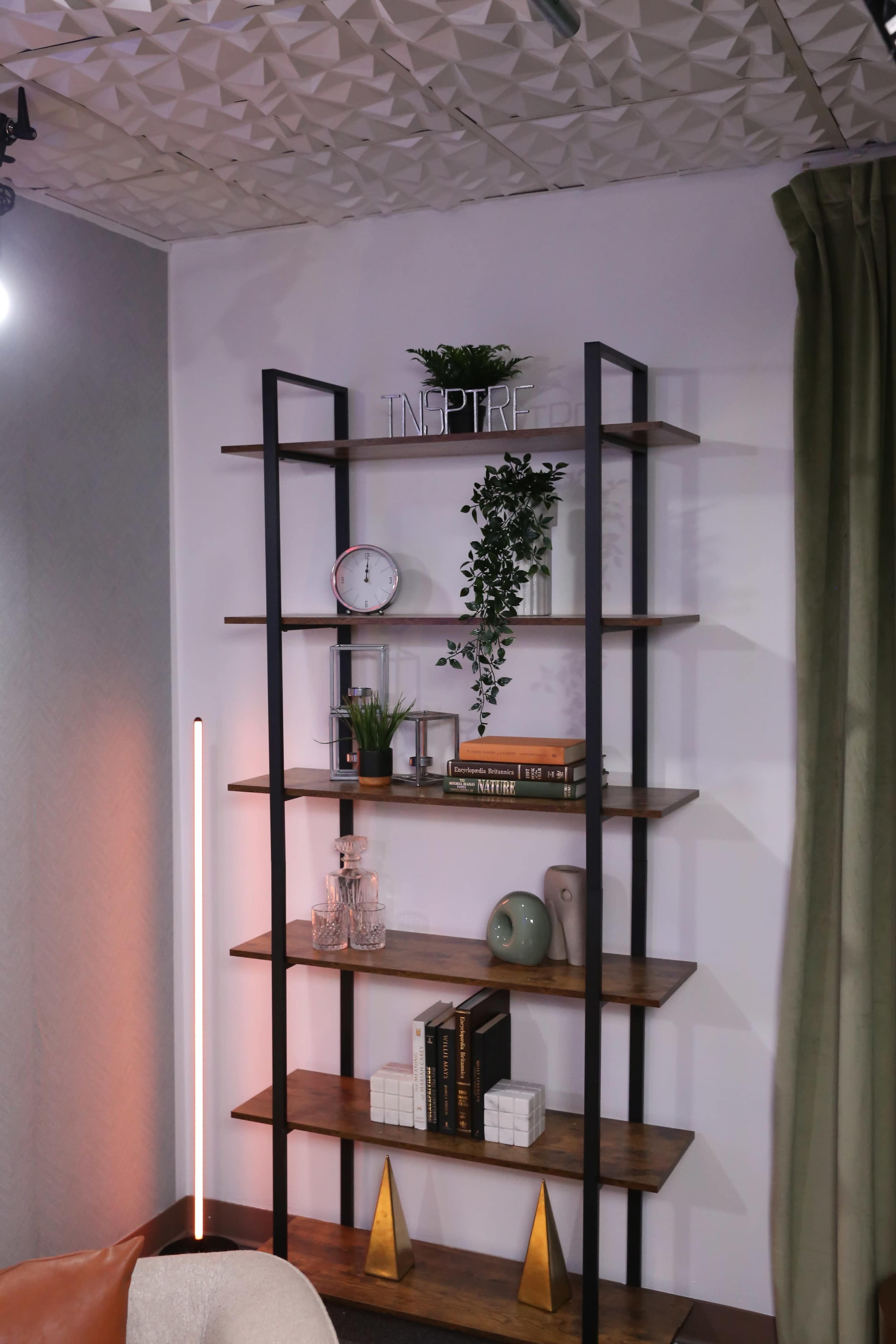 A metal and wood bookshelf displays various decorative items, plants, and books against a textured wall and curtain.