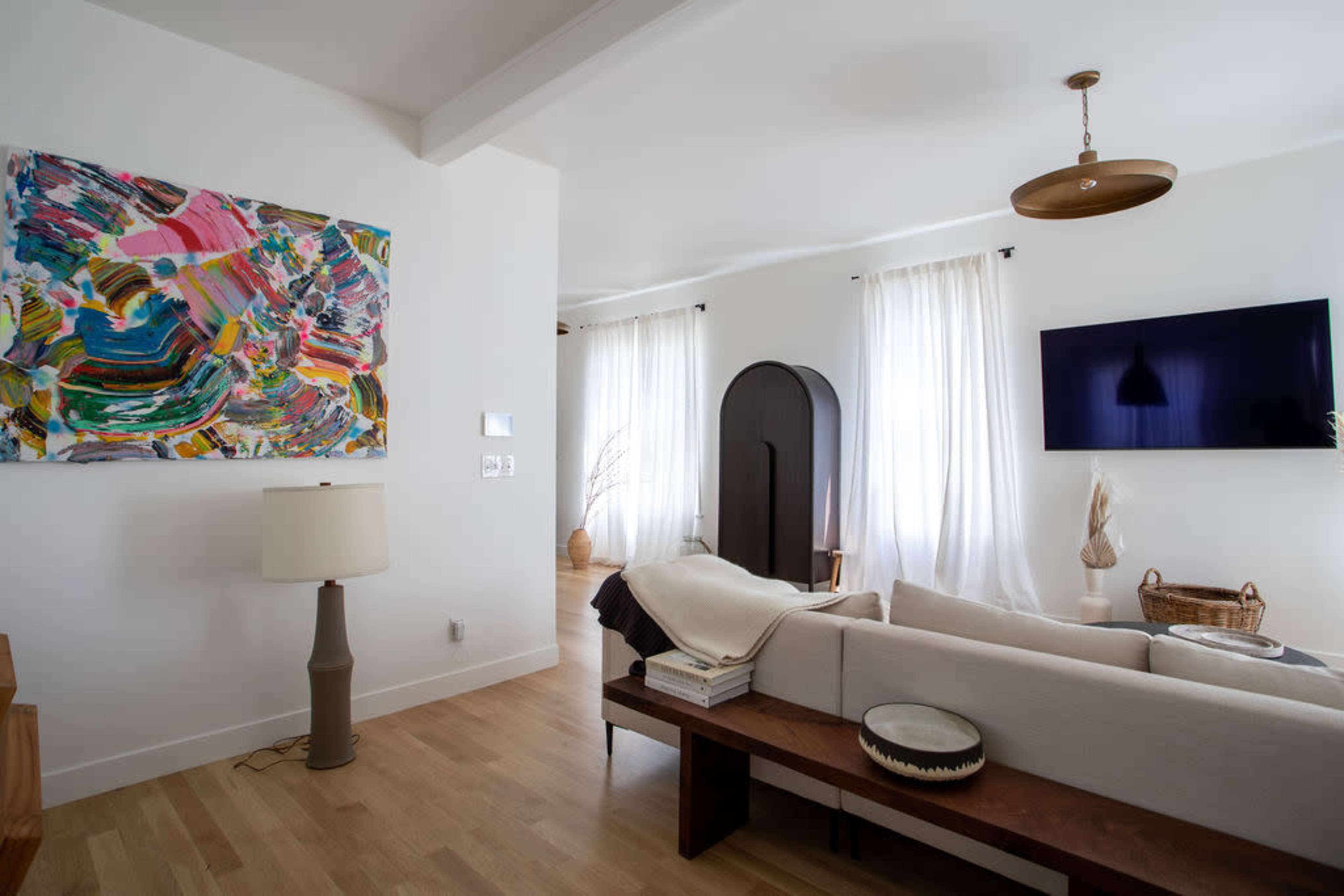 A bright living room features a colorful abstract painting, a modern lamp, and a television mounted on the wall, with muted furnishings and large windows allowing natural light.