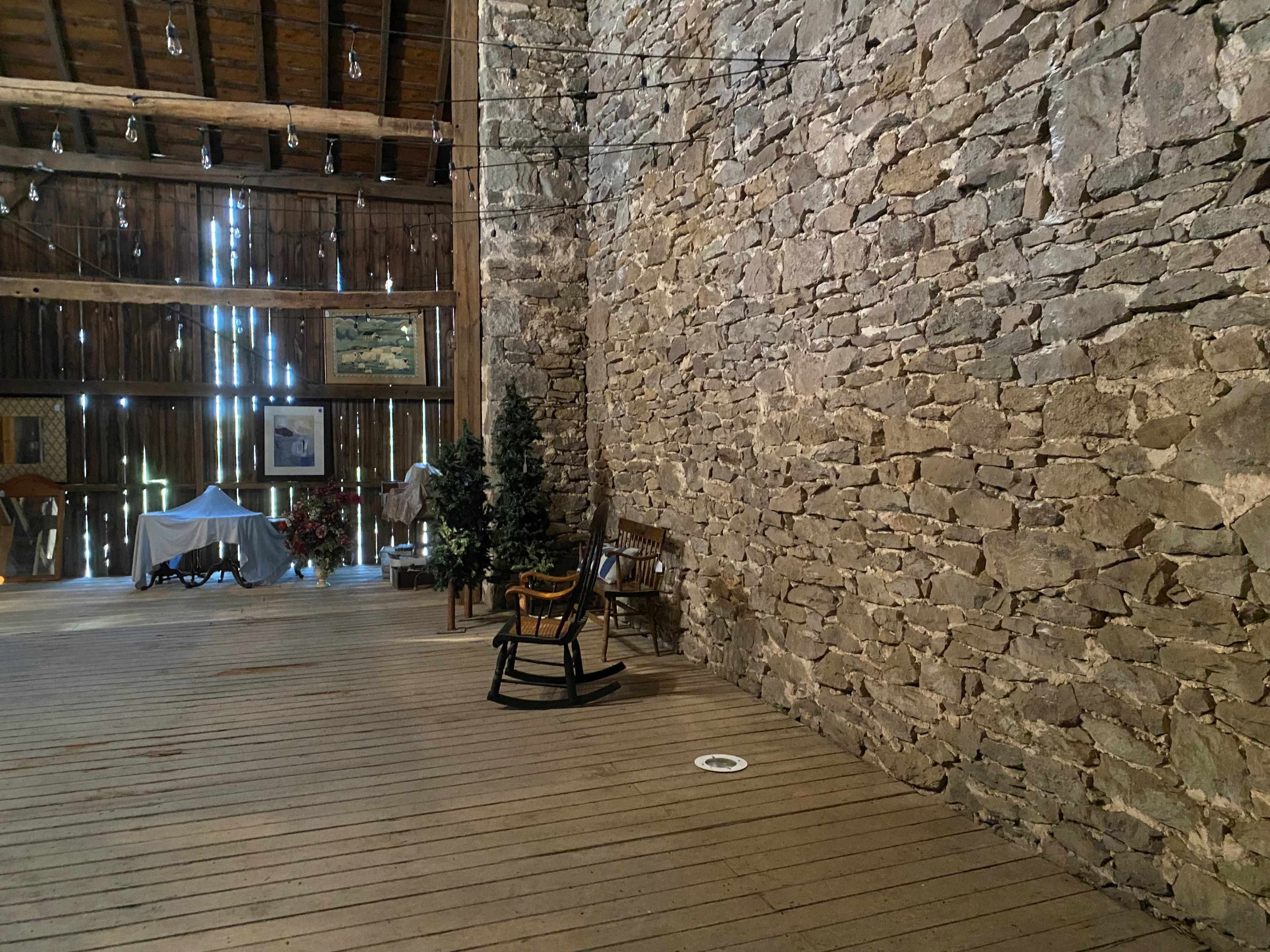 The interior of a rustic barn with a stone wall, wooden floors, and various pieces of furniture, including a rocking chair and a Christmas tree.