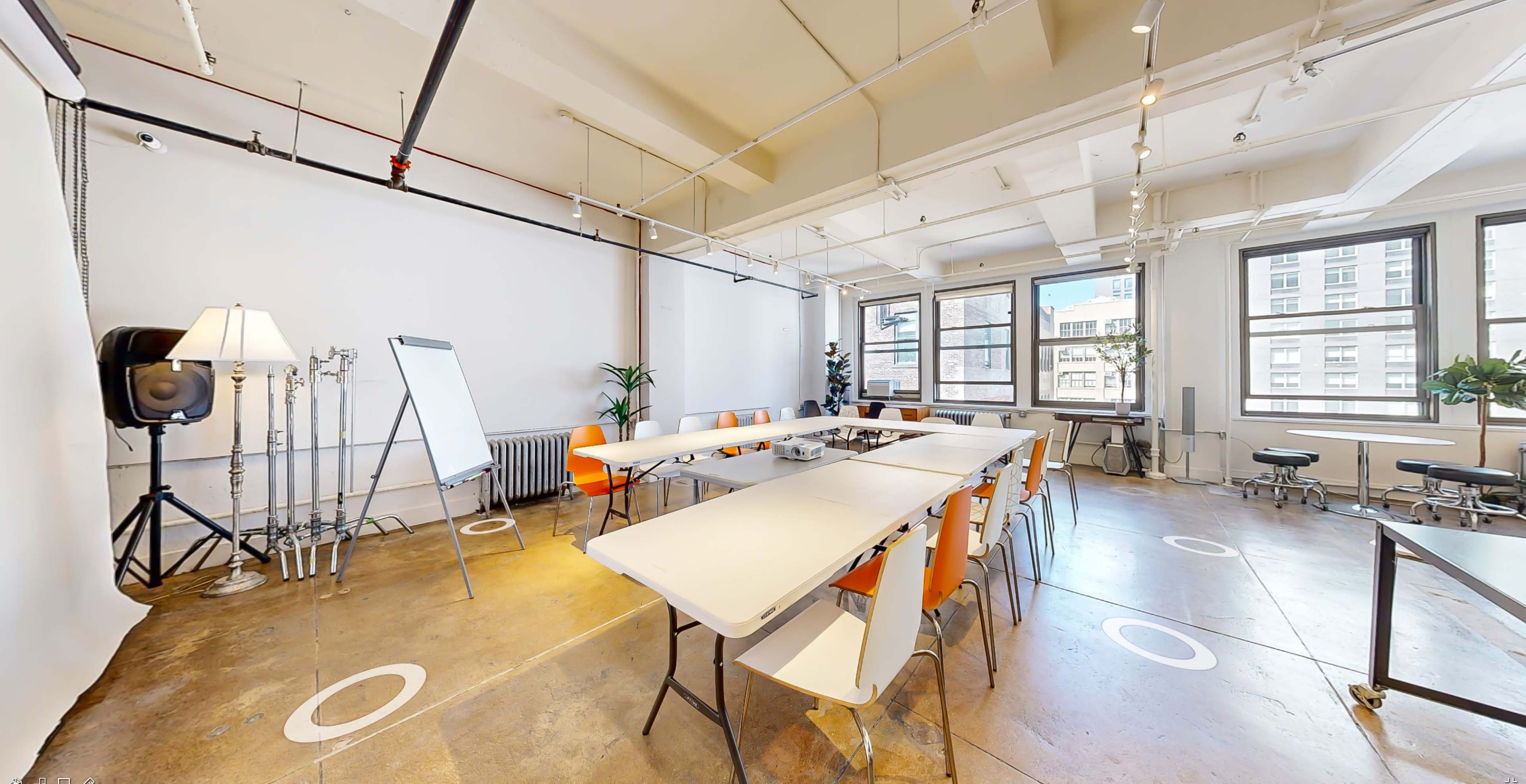 The image shows a modern meeting room with several white tables and orange chairs arranged in a spacious layout, along with large windows letting in natural light.