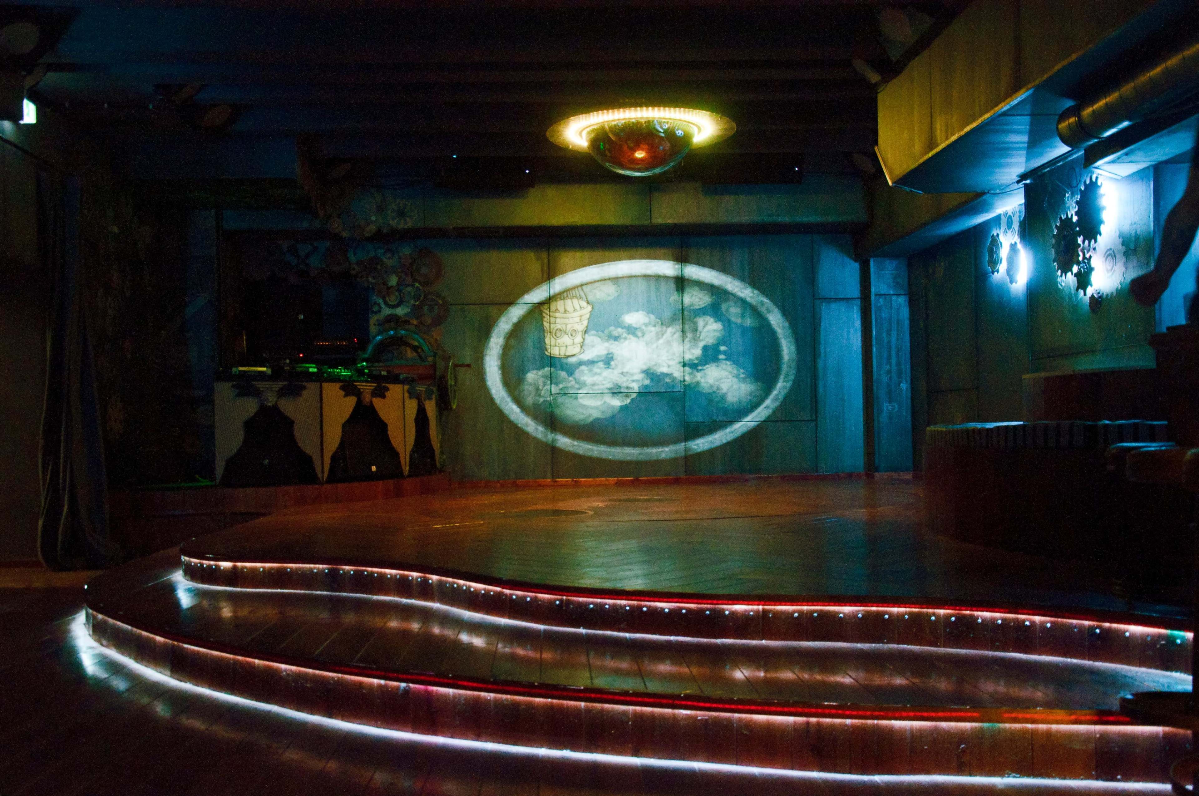The image shows a dimly lit nightclub stage with a circular projected design on the wall and illuminated steps leading up to it.