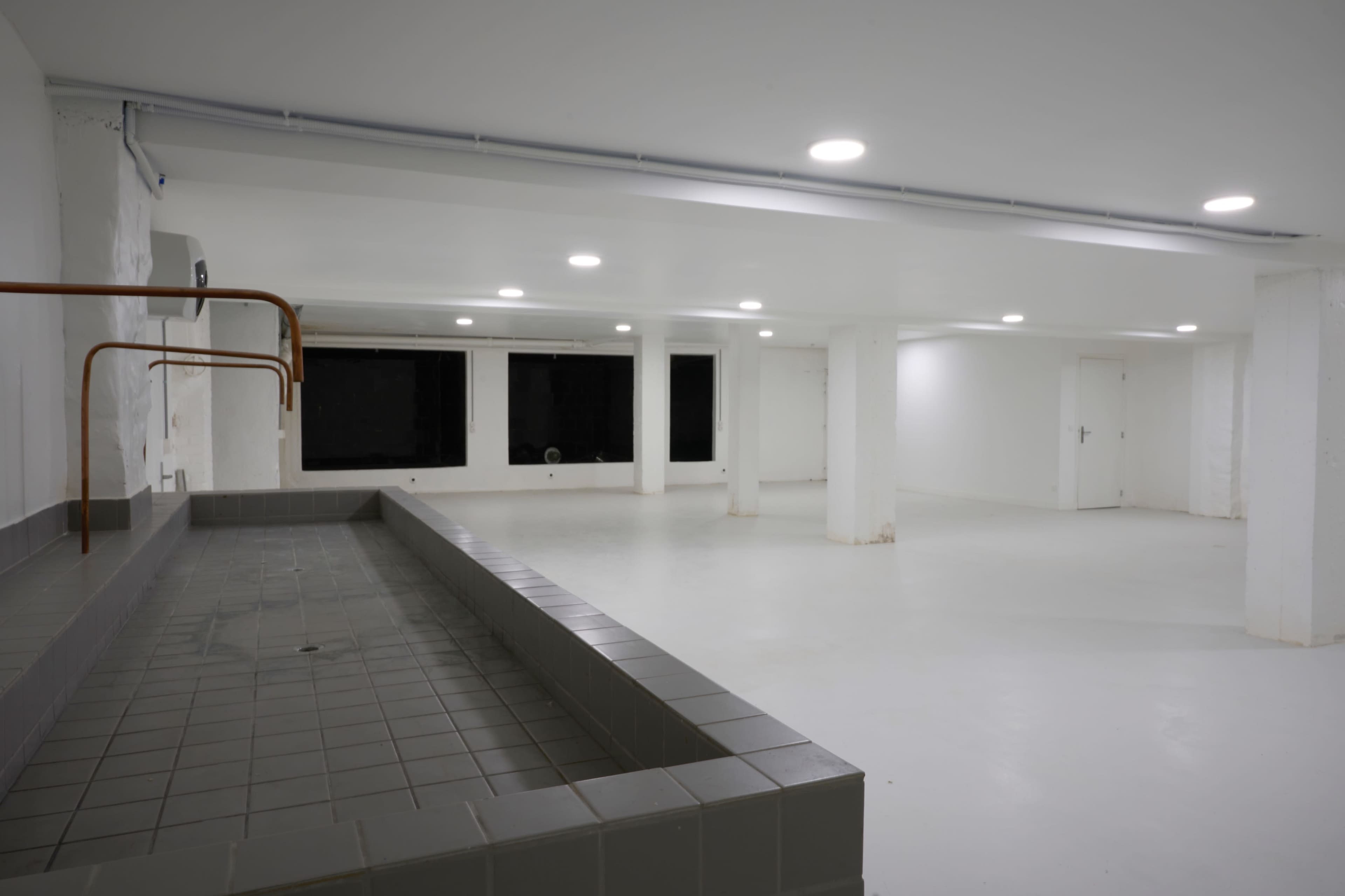 A large, empty room with white walls, gray tiled flooring, and a tiled seating area along one side, illuminated by ceiling lights.
