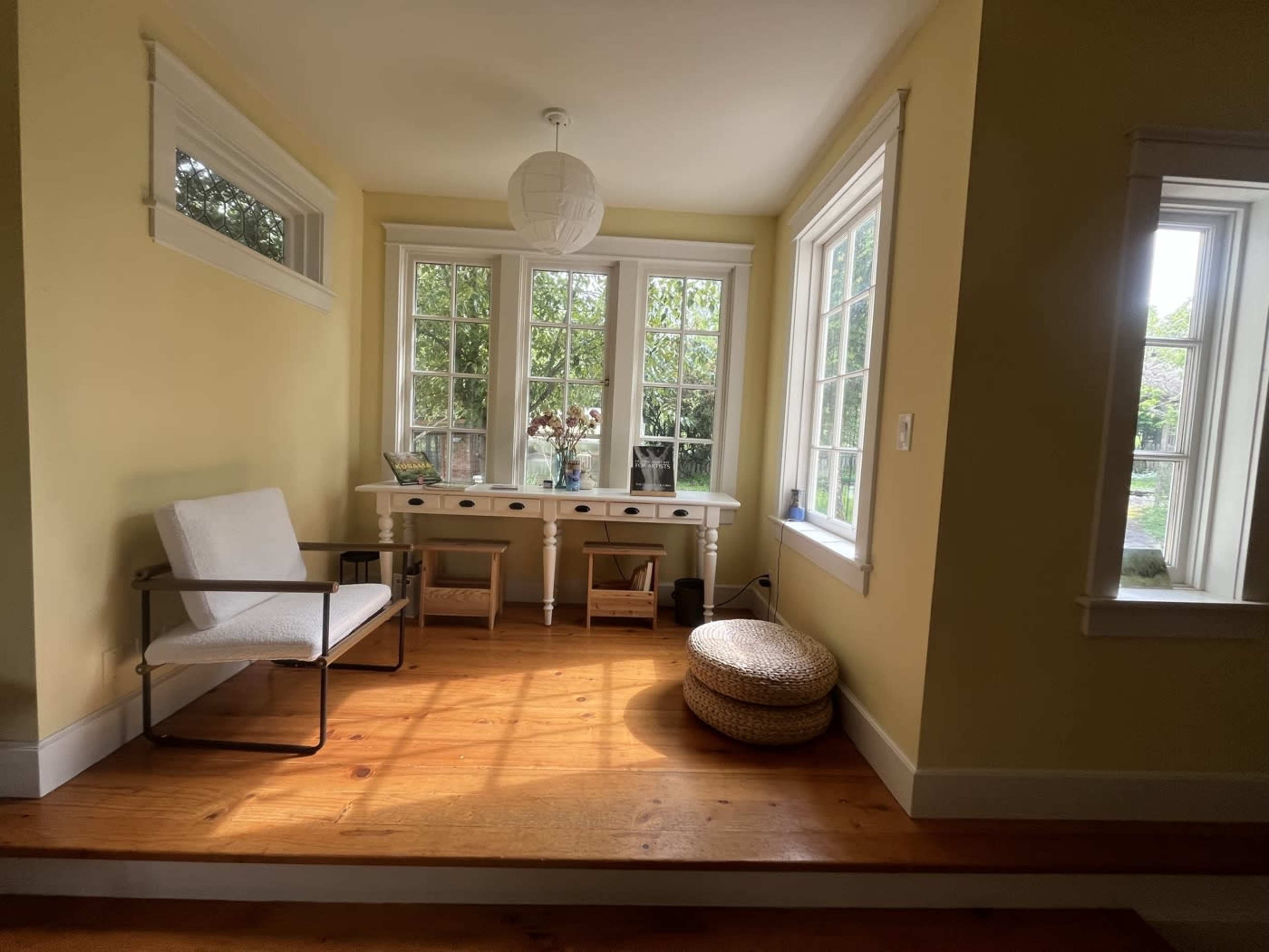 The image shows a brightly lit room with large windows, a small wooden table, a white chair, and a round woven ottoman on the floor.