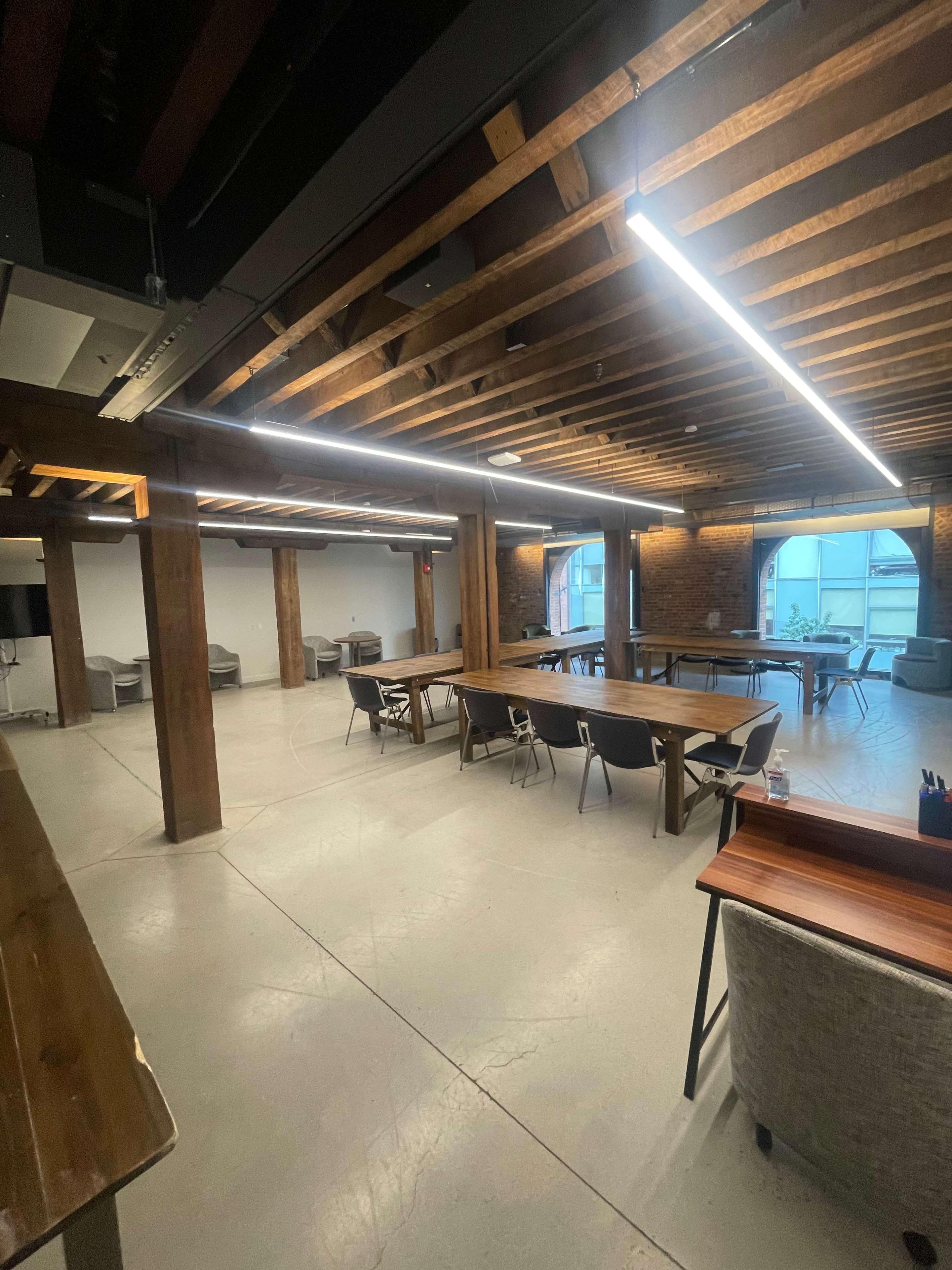 A spacious interior of a modern office with wooden beams, large windows, and multiple tables and chairs arranged throughout.