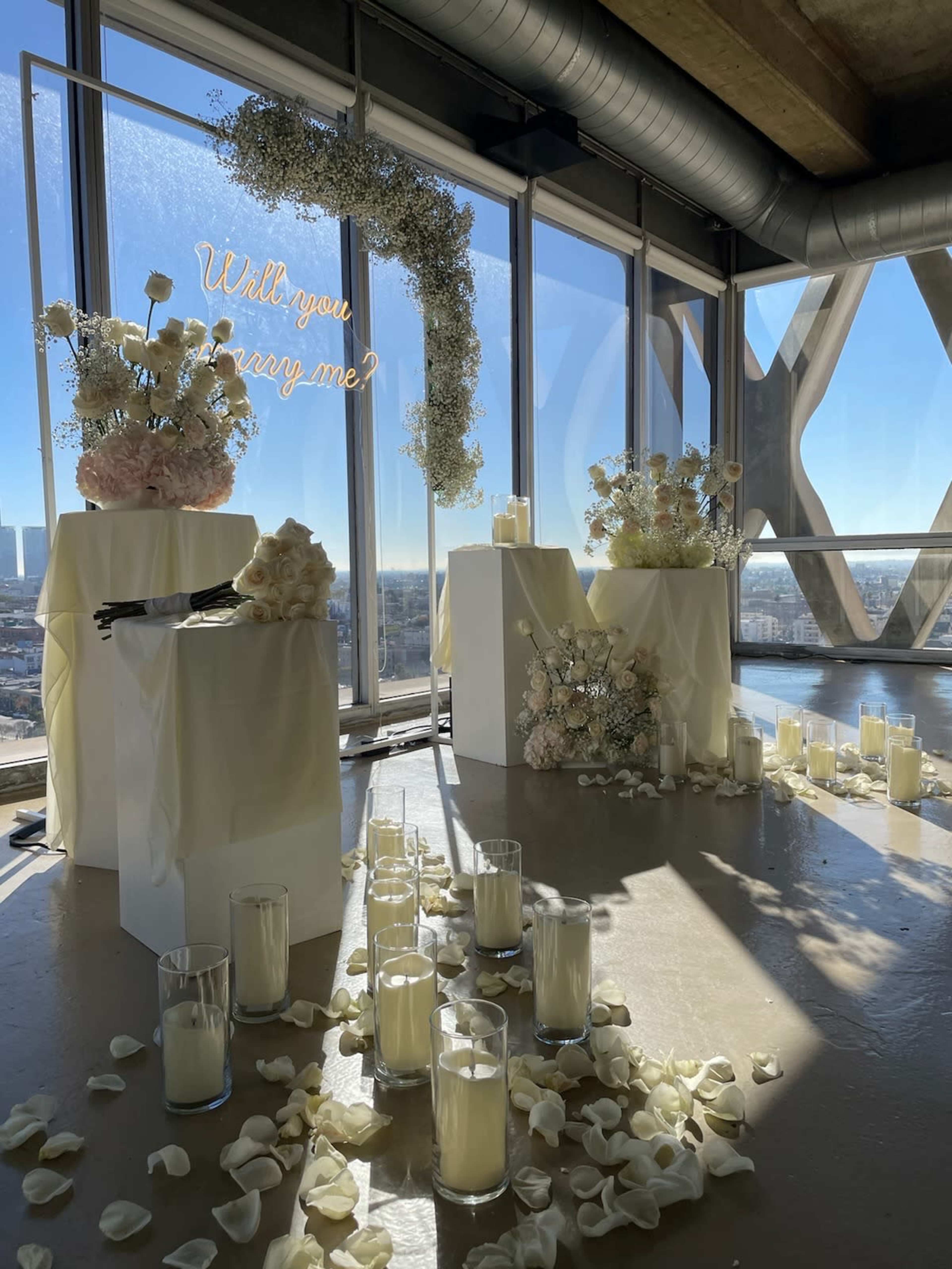 A beautifully arranged engagement setup features white floral arrangements and candles scattered among rose petals, all set against a backdrop of large windows showcasing a city view.