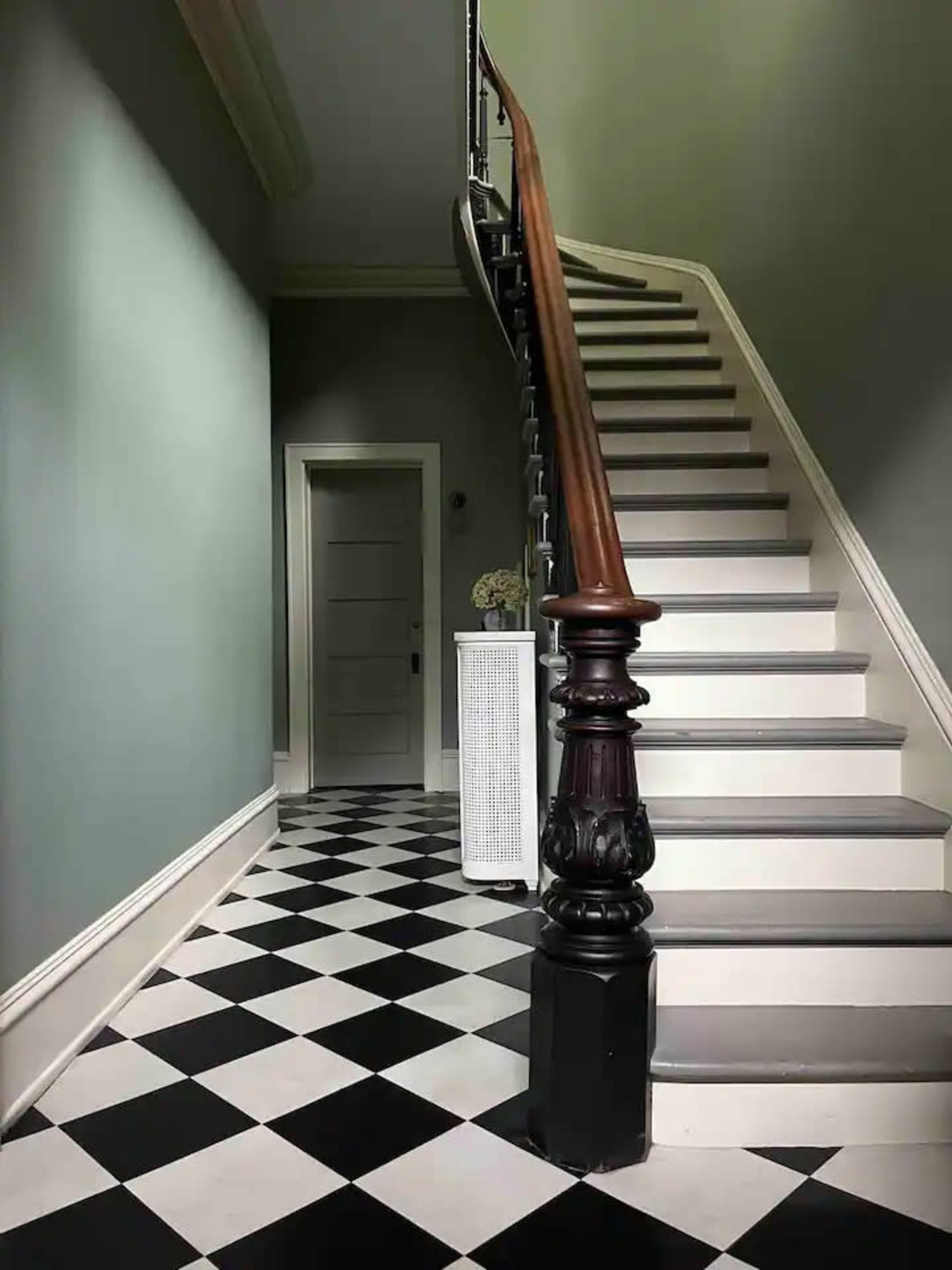 A staircase with a wooden banister, leading to an upper floor, alongside a checkered black and white floor and a closed door in the hallway.