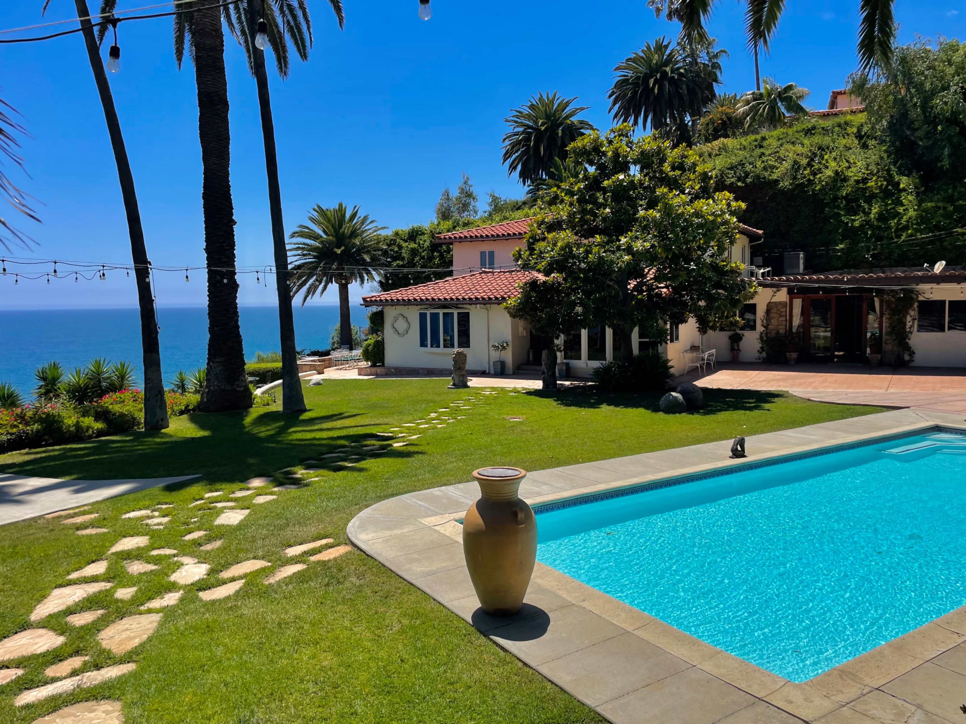 A spacious backyard features a swimming pool, surrounded by palm trees and a clear view of the ocean, with a house in the background.