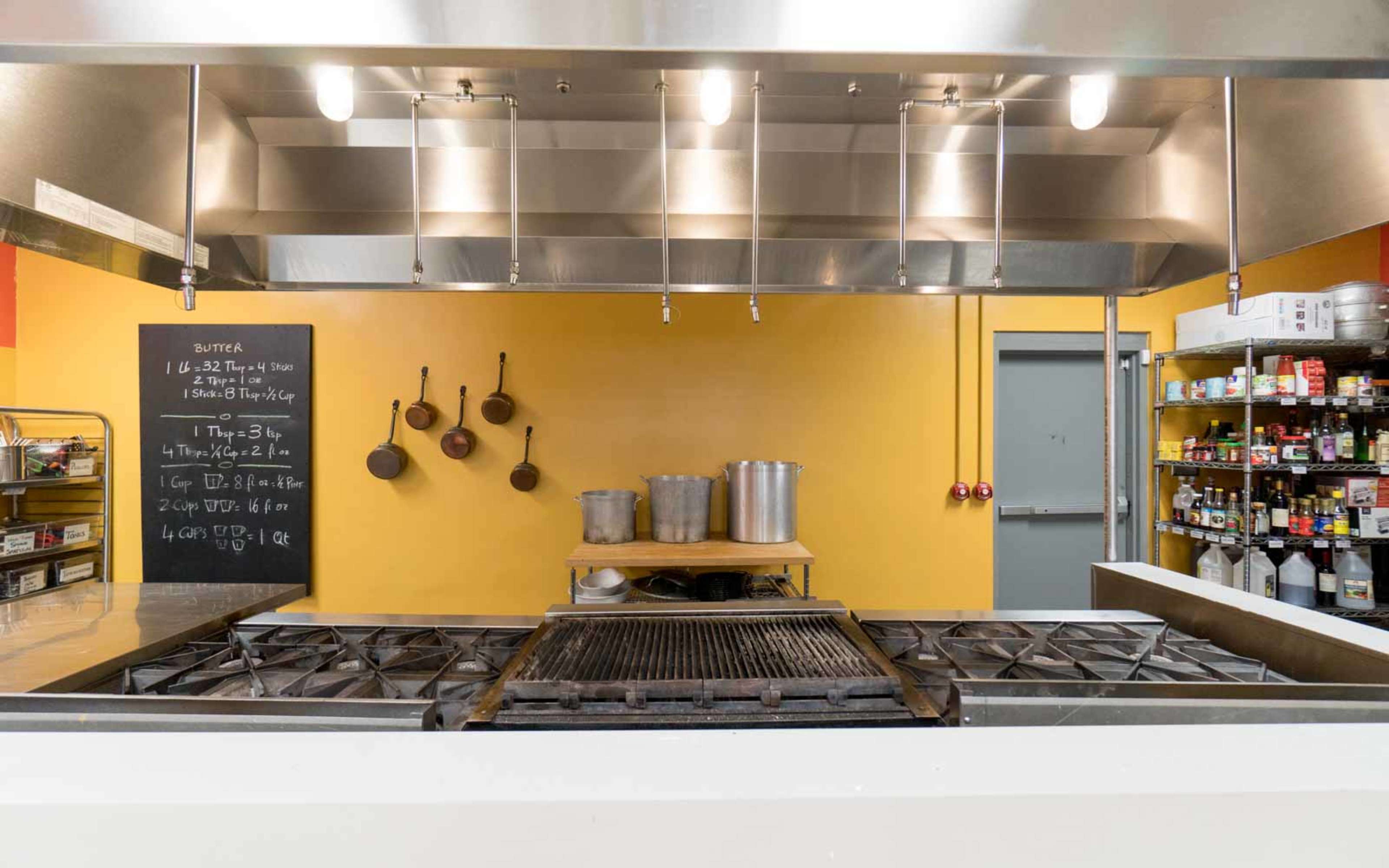 A commercial kitchen with a gas stove, hanging cookware, a chalkboard with recipes, and shelves stocked with various cooking ingredients.