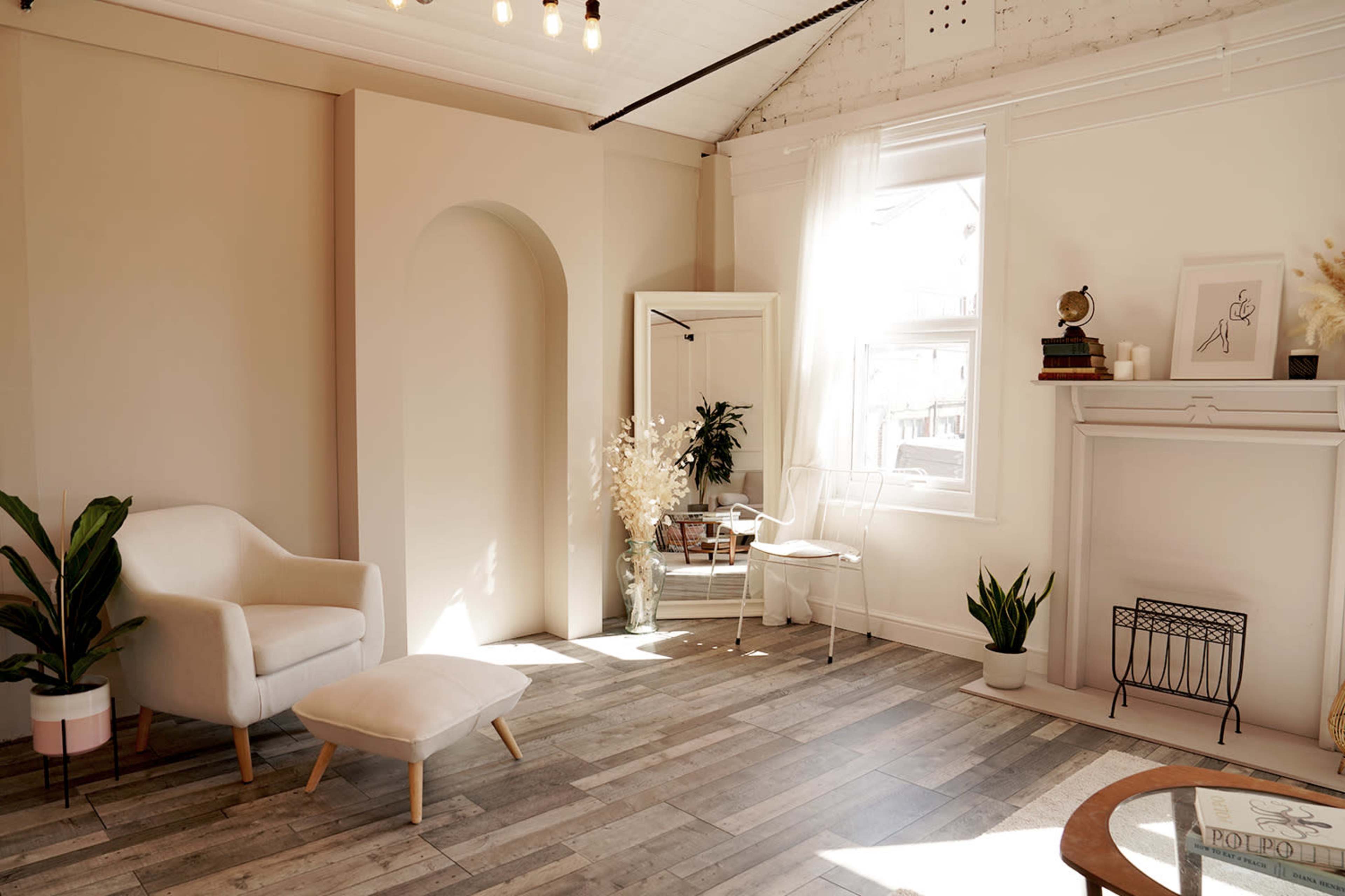 The image shows a bright and minimalistic interior space featuring a white armchair, a small ottoman, a tall mirror, and various plants, with a decorative fireplace and natural light streaming in through the windows.