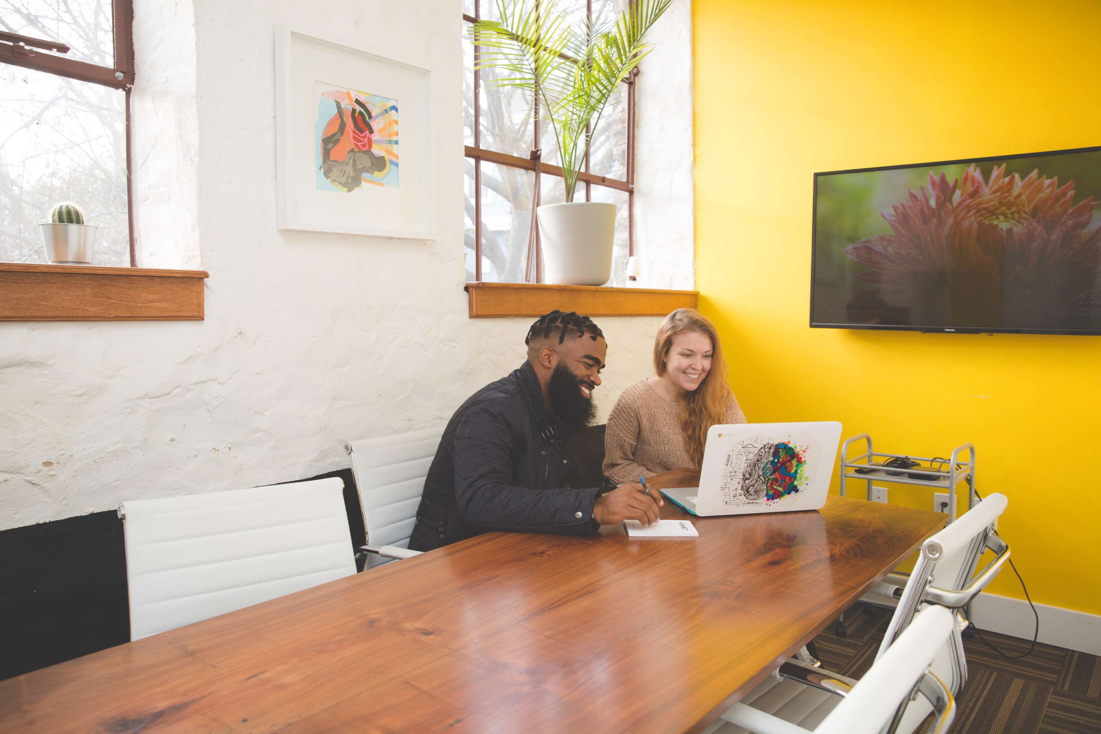 A man and a woman are seated at a wooden conference table, working on a laptop in a bright room with yellow walls and large windows.