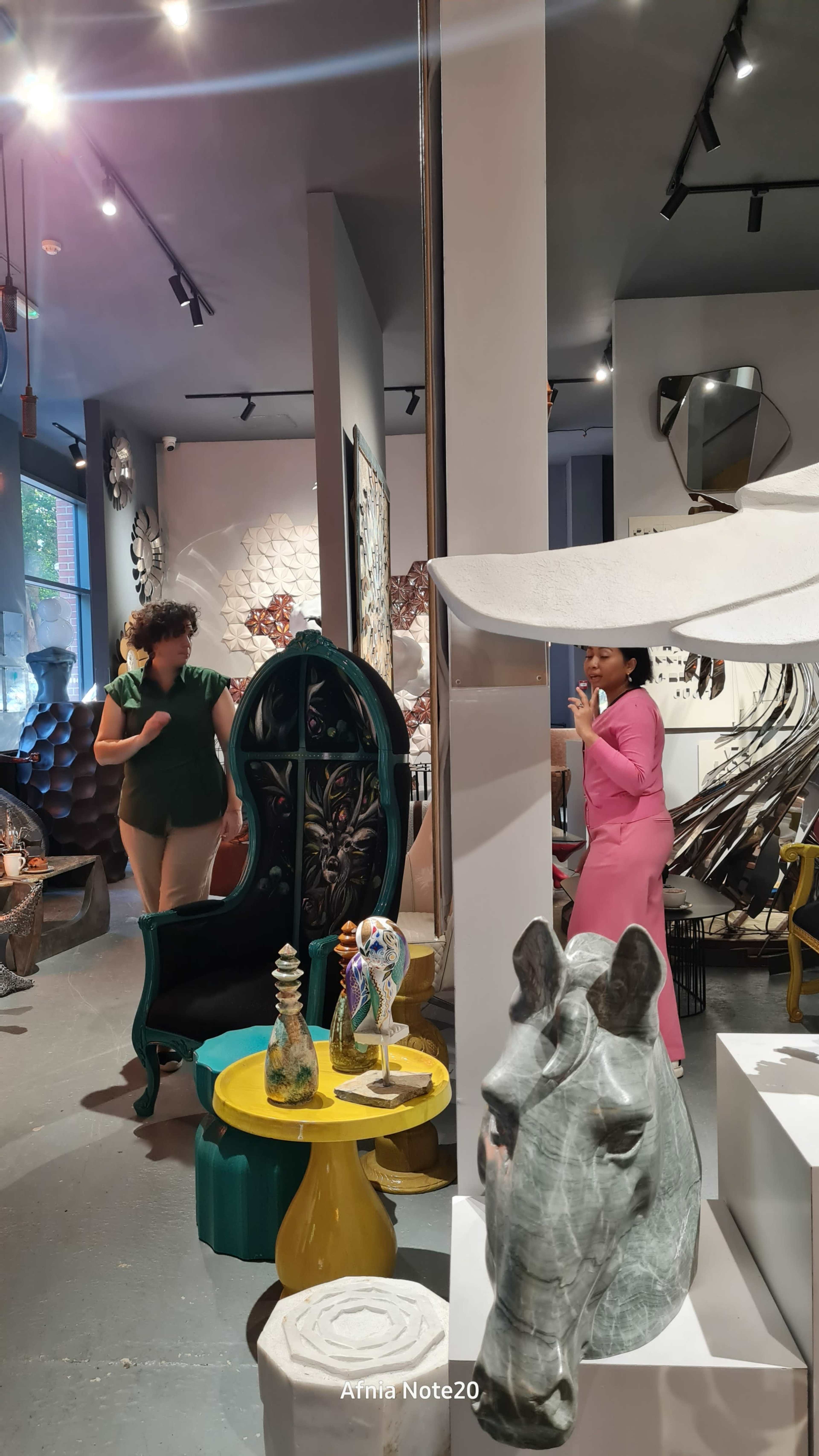 The image shows an eclectic furniture store filled with unique decorative pieces, including a large green chair, colorful sculptures, and a horse statue, with two women engaged in conversation among the displays.