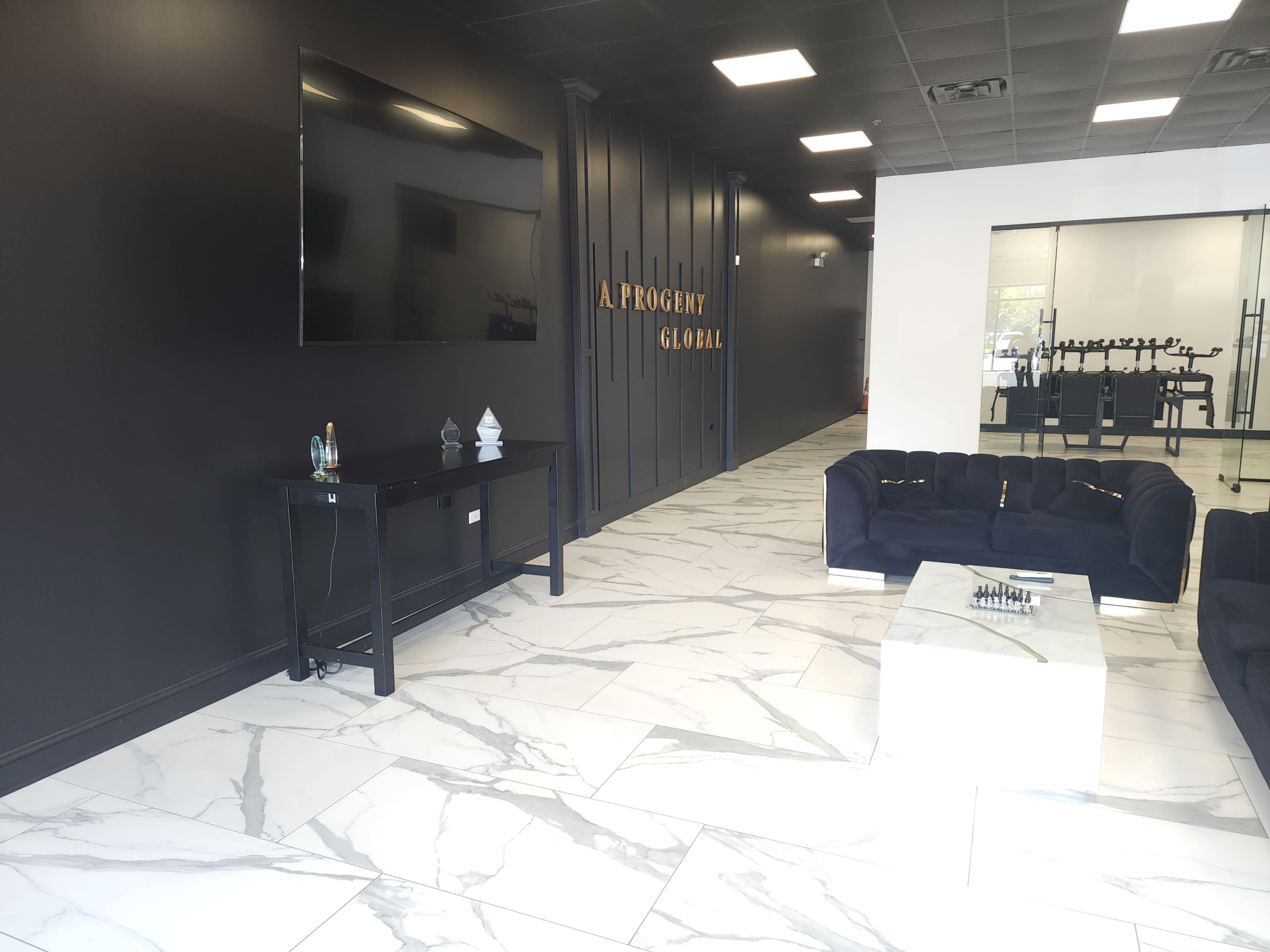 The image shows a modern office reception area with dark walls, marble flooring, a large TV mounted on the wall, a black sofa, and a glass coffee table.