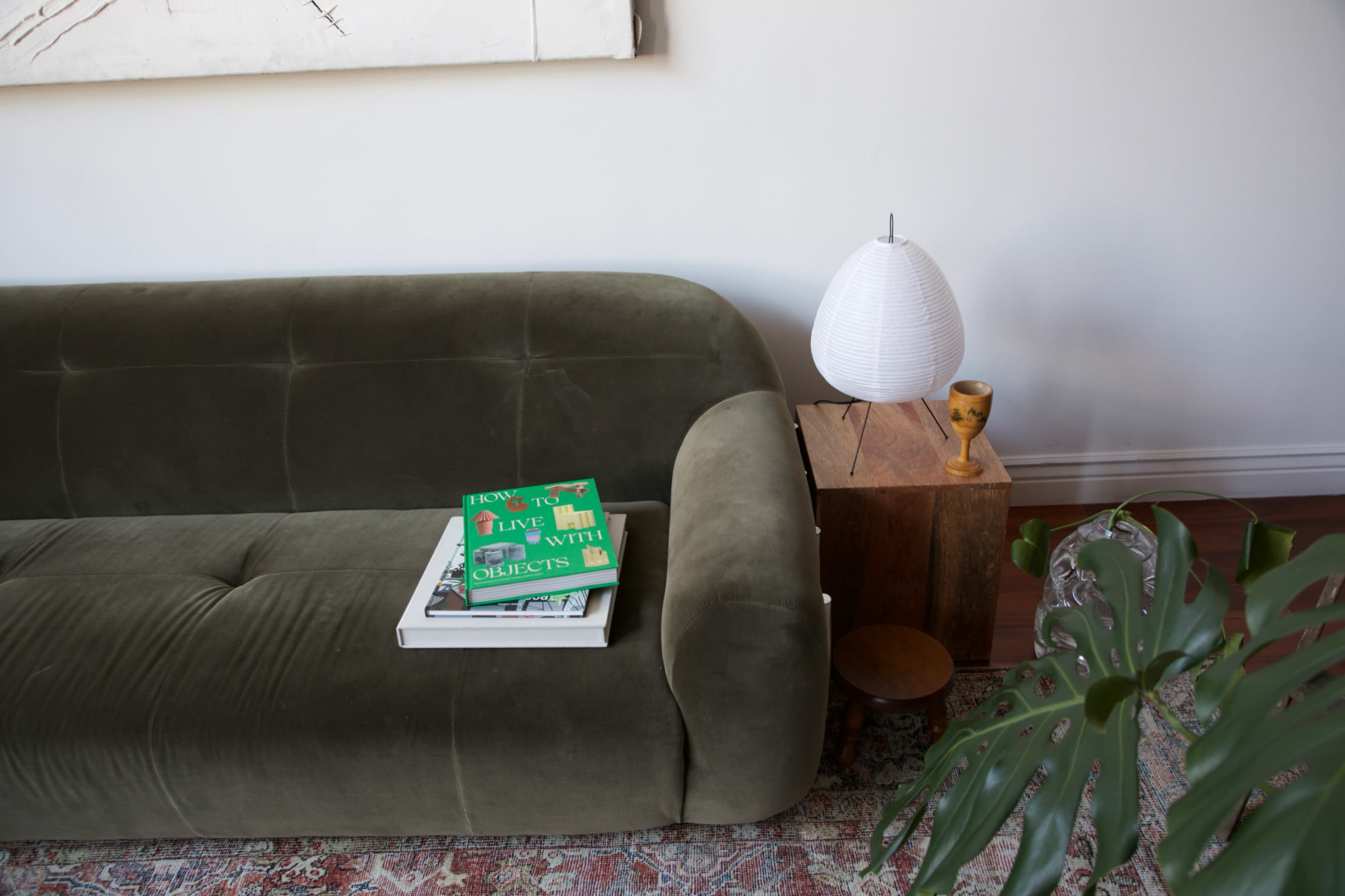 A green sofa with a book and a magazine on it is positioned next to a wooden side table featuring a sculptural lamp and a small decorative piece.