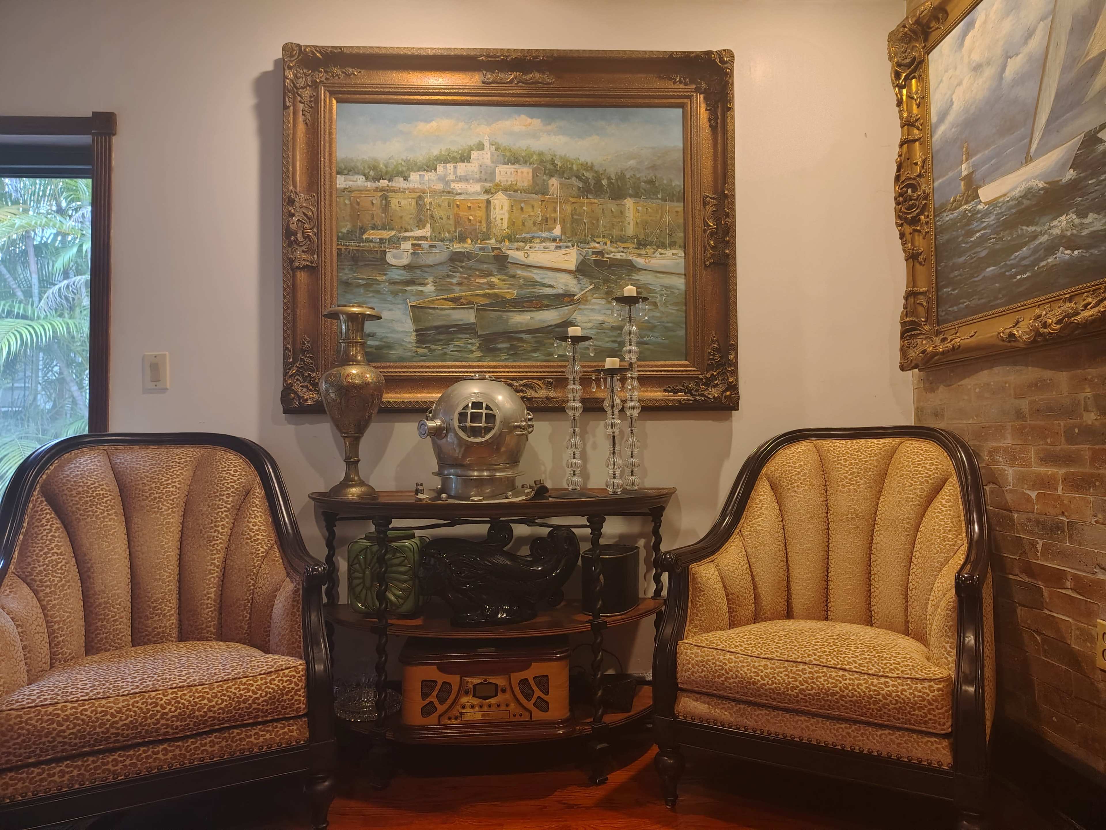 The image shows two patterned armchairs flanking a small table adorned with decorative items, including a vintage diving helmet and a painting of a coastal scene on the wall above.