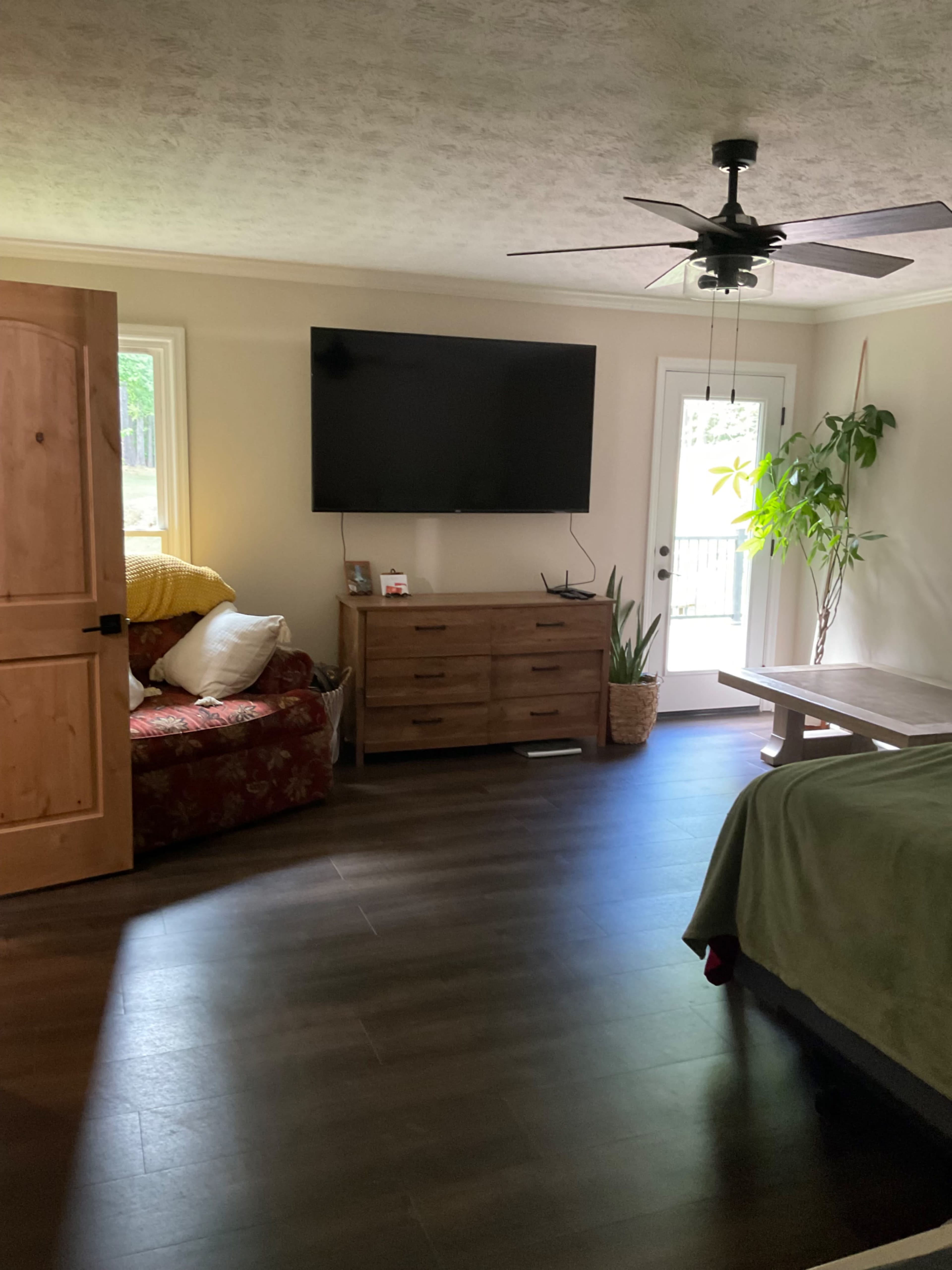 A spacious room features a brown sofa, a wooden dresser, a large television mounted on the wall, and a door leading to an outdoor area.