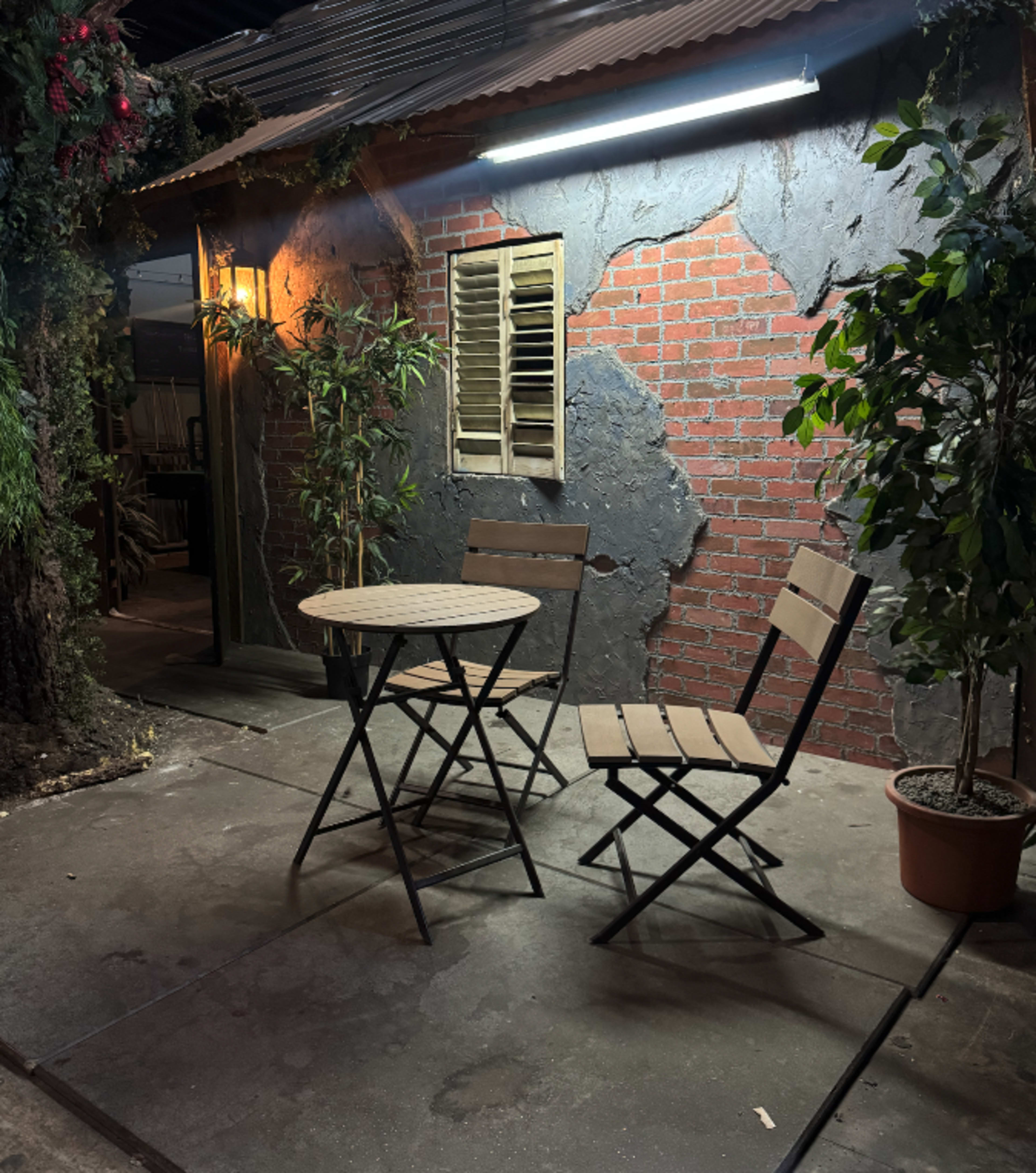 The image shows a small outdoor seating area with two folding chairs and a round table beside a rustic-looking wall featuring bricks and a window with shutters.