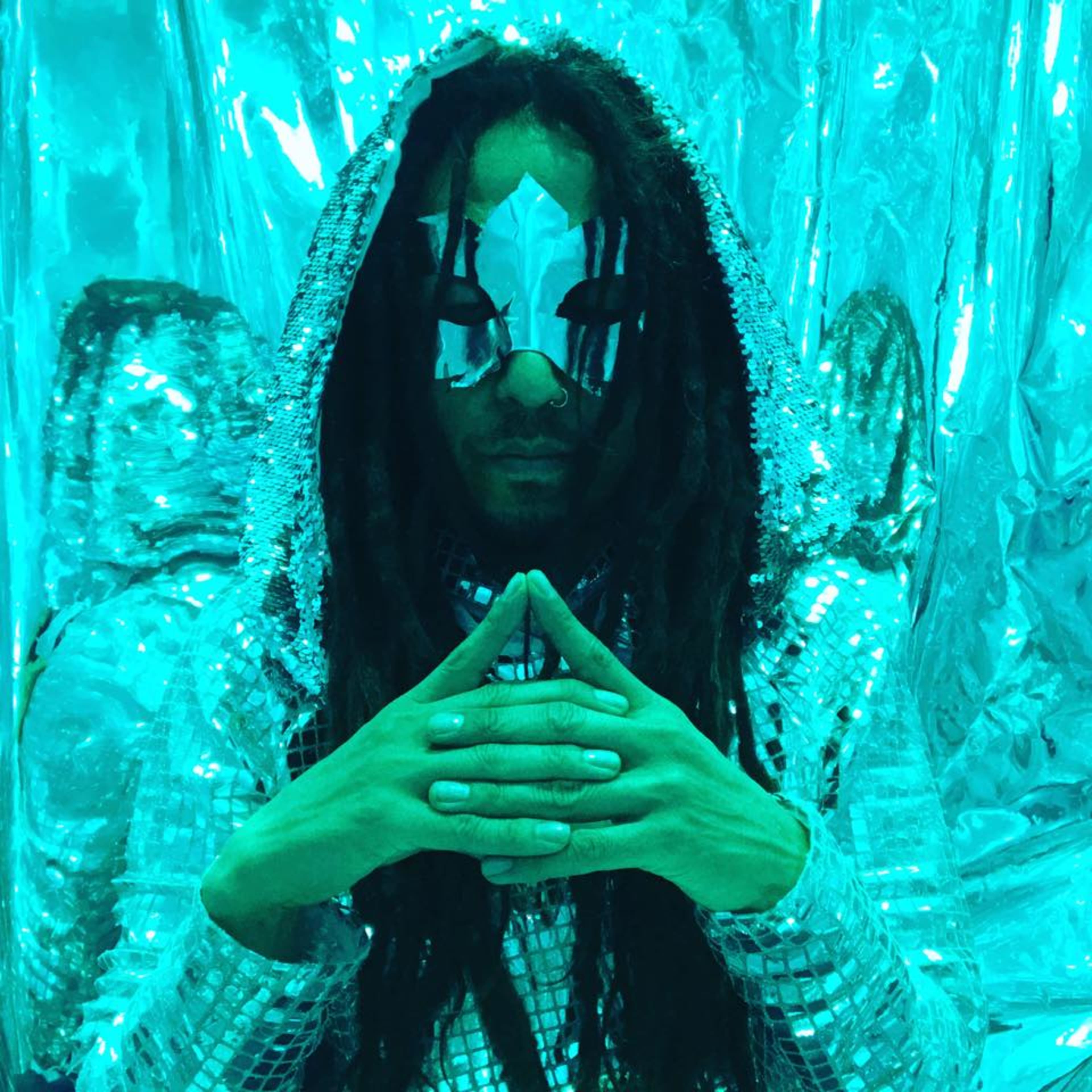 A person wearing a silver costume and a decorative mask sits with hands clasped in front of a shimmering turquoise background.
