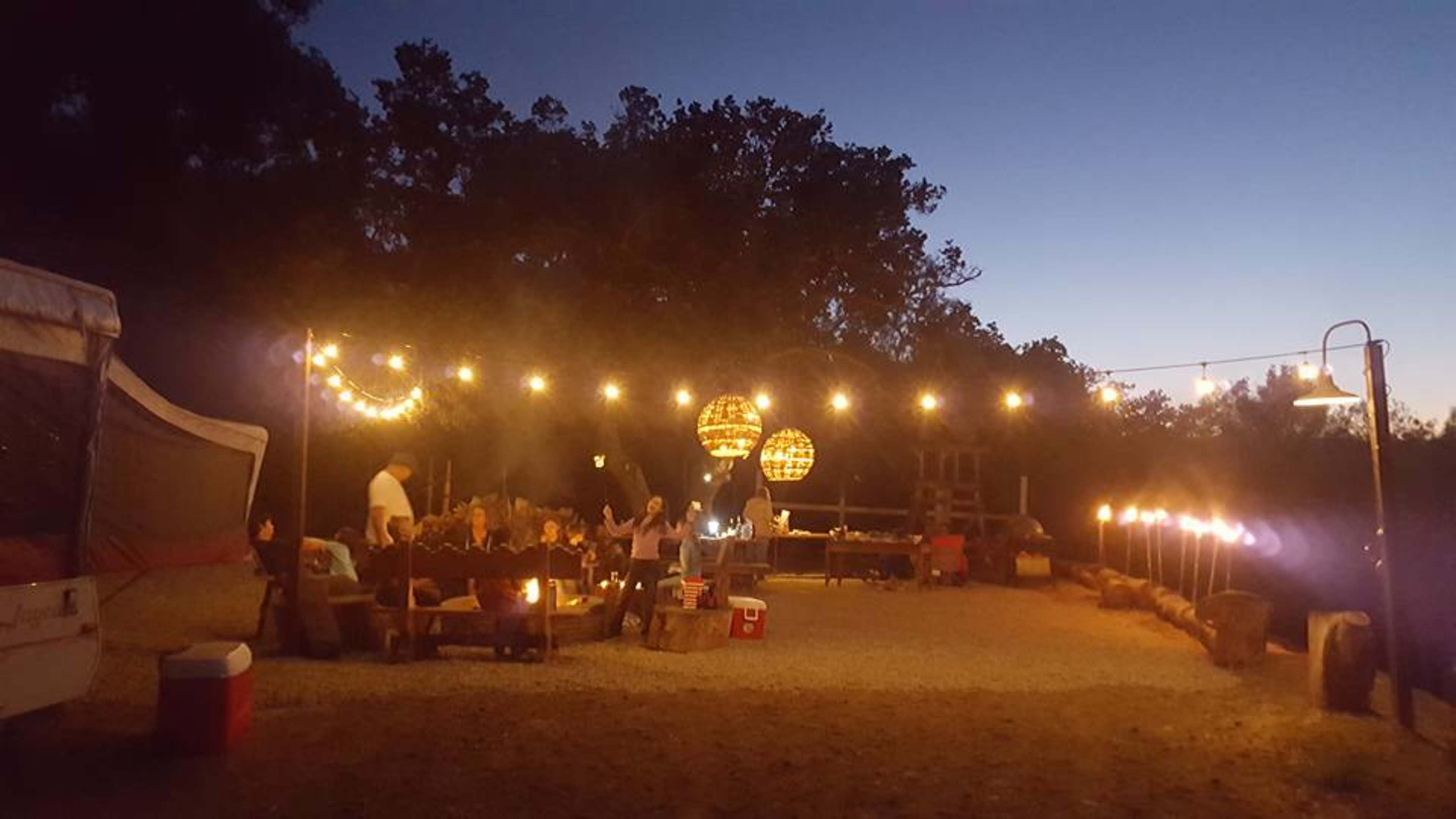 A camping area is illuminated by string lights and lanterns, featuring a seating arrangement around a fire pit.
