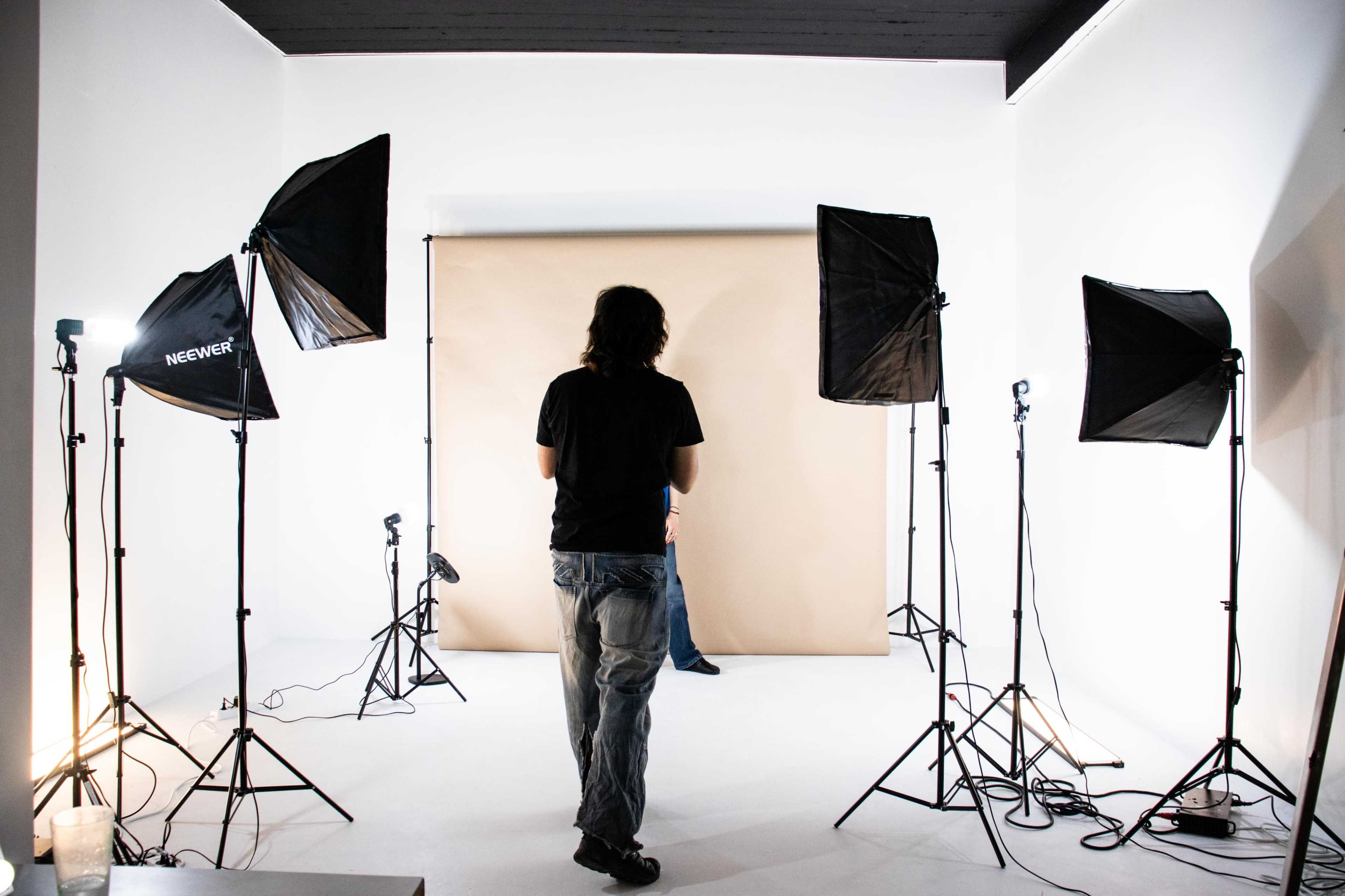 A person stands in a photography studio surrounded by softbox lights and a beige backdrop.