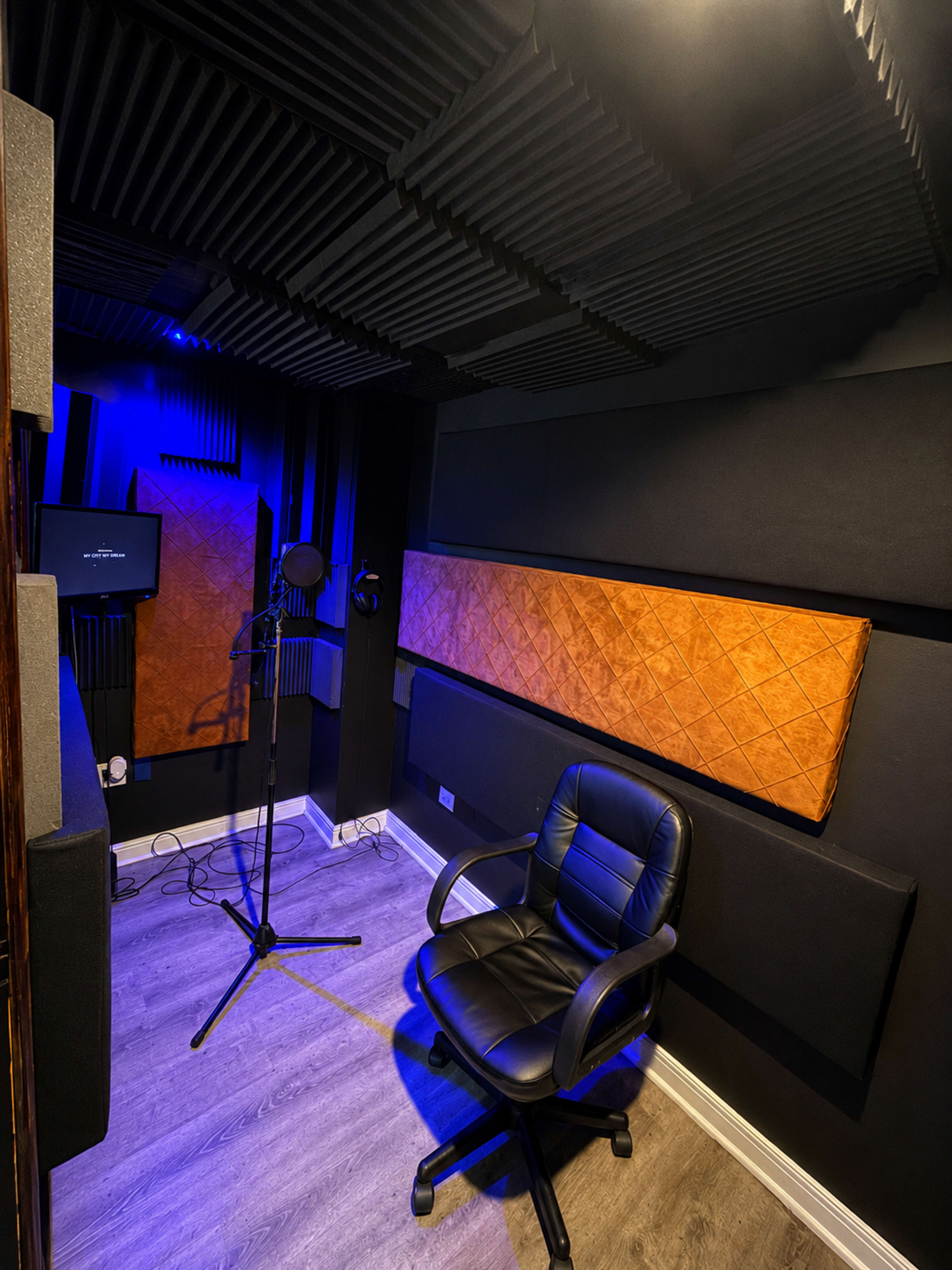 The image shows a small soundproof recording studio with black acoustic panels, a microphone on a stand, and a black leather chair.