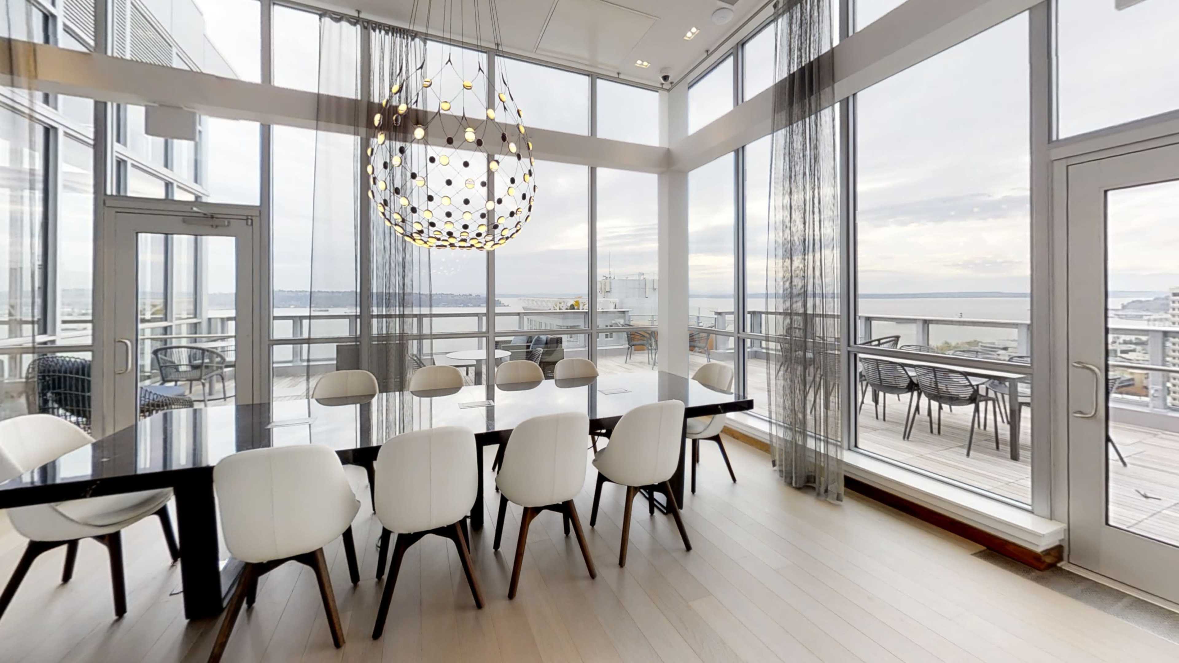 A modern dining area with a large table surrounded by white chairs, featuring large windows that overlook a waterfront view.