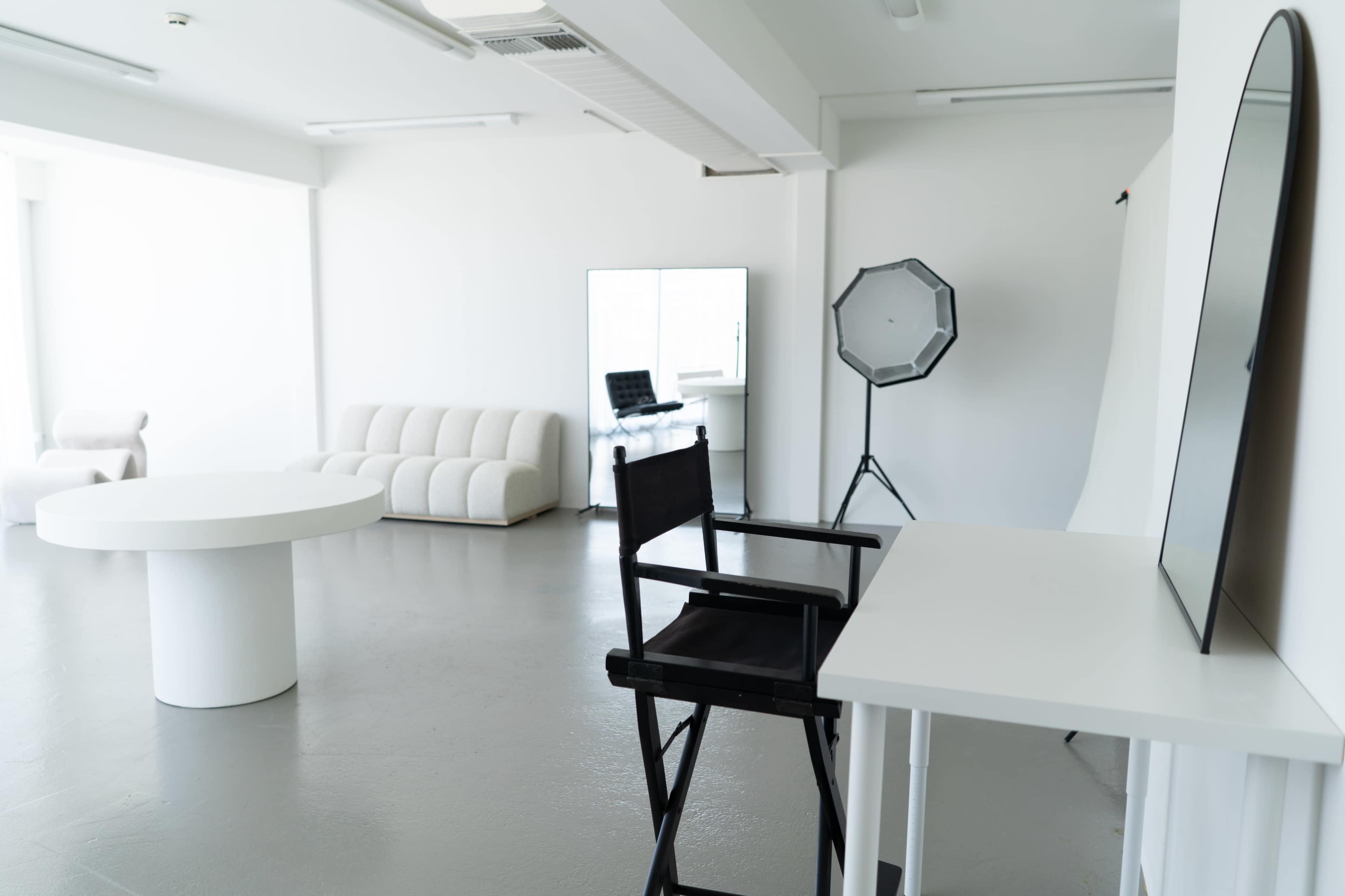 The image shows a spacious, minimalistic studio featuring a round table, a black director's chair, a large mirror, a soft couch, and professional lighting equipment.