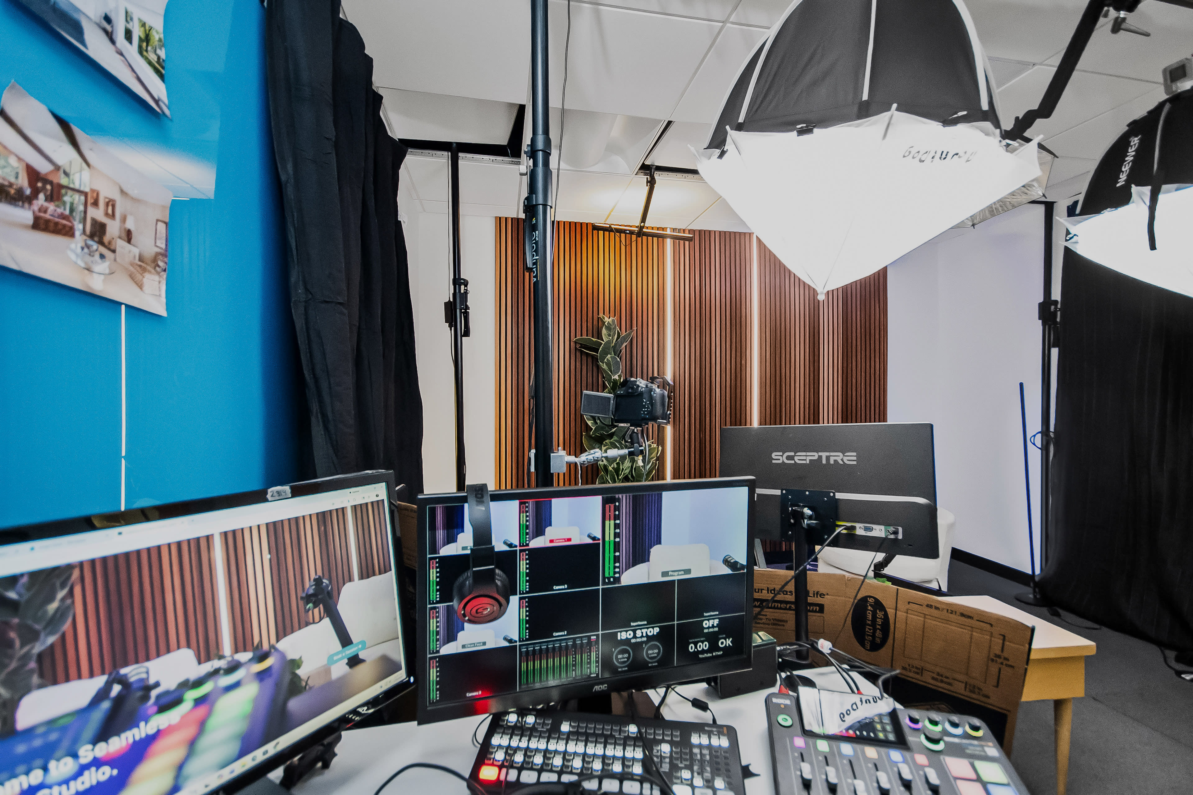 The image shows a video production setup with multiple monitors displaying software, a camera on a tripod, and professional lighting equipment in a studio environment.