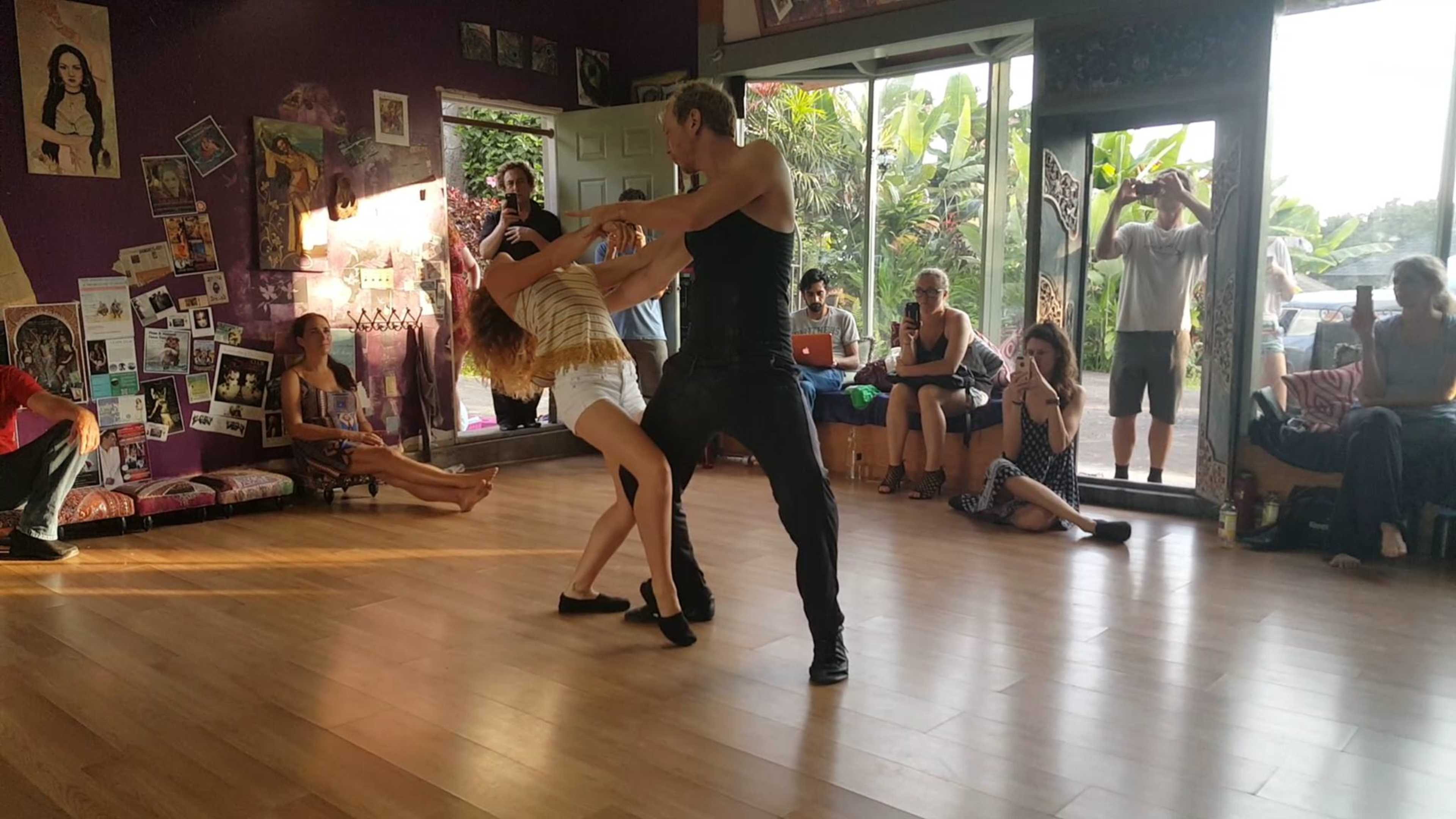 Two dancers perform a routine in a studio with spectators observing from various spots around the room.