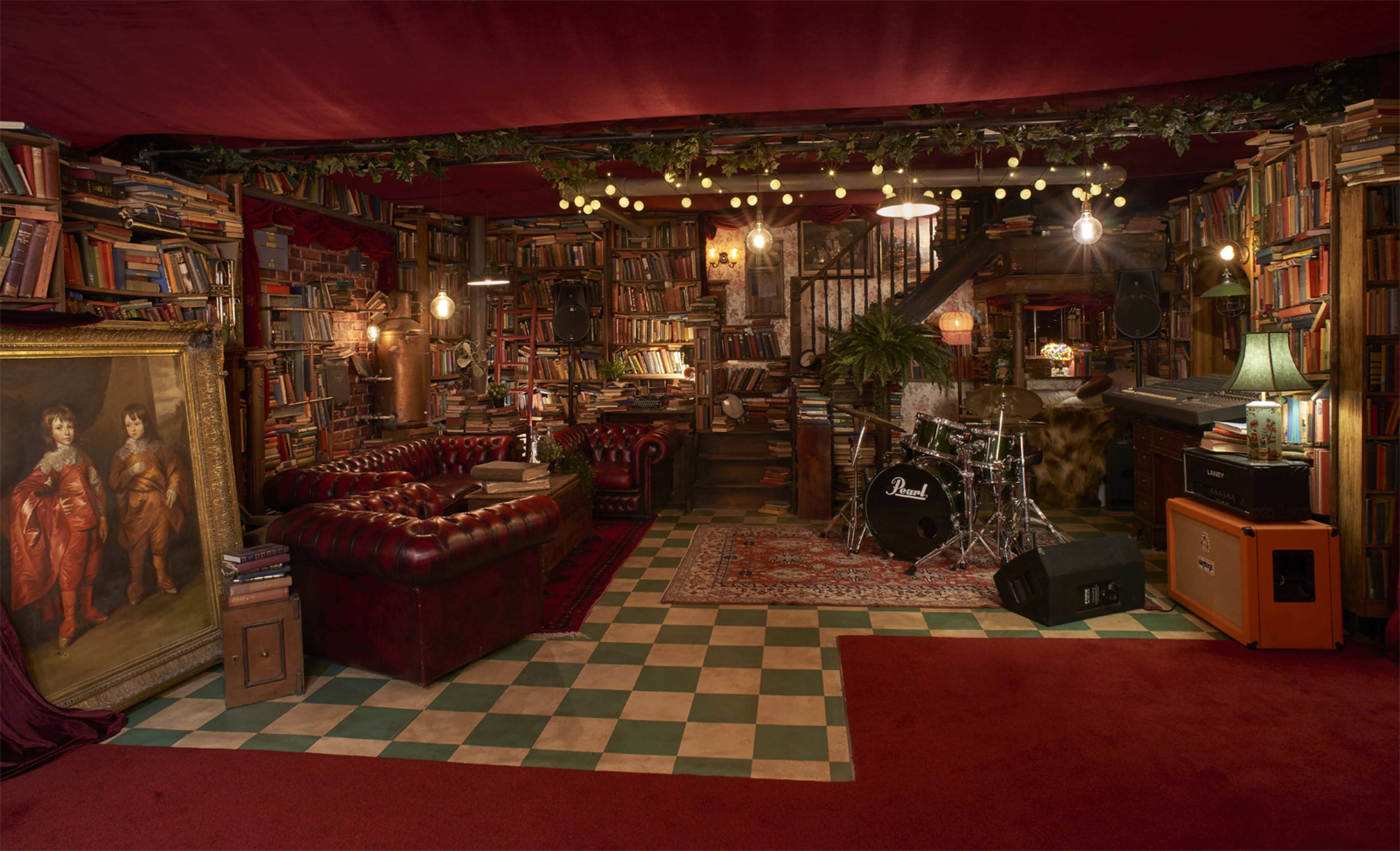 The image shows a cozy room filled with bookshelves, a red leather couch, a drum set, and decorative string lights, creating a warm, inviting atmosphere.