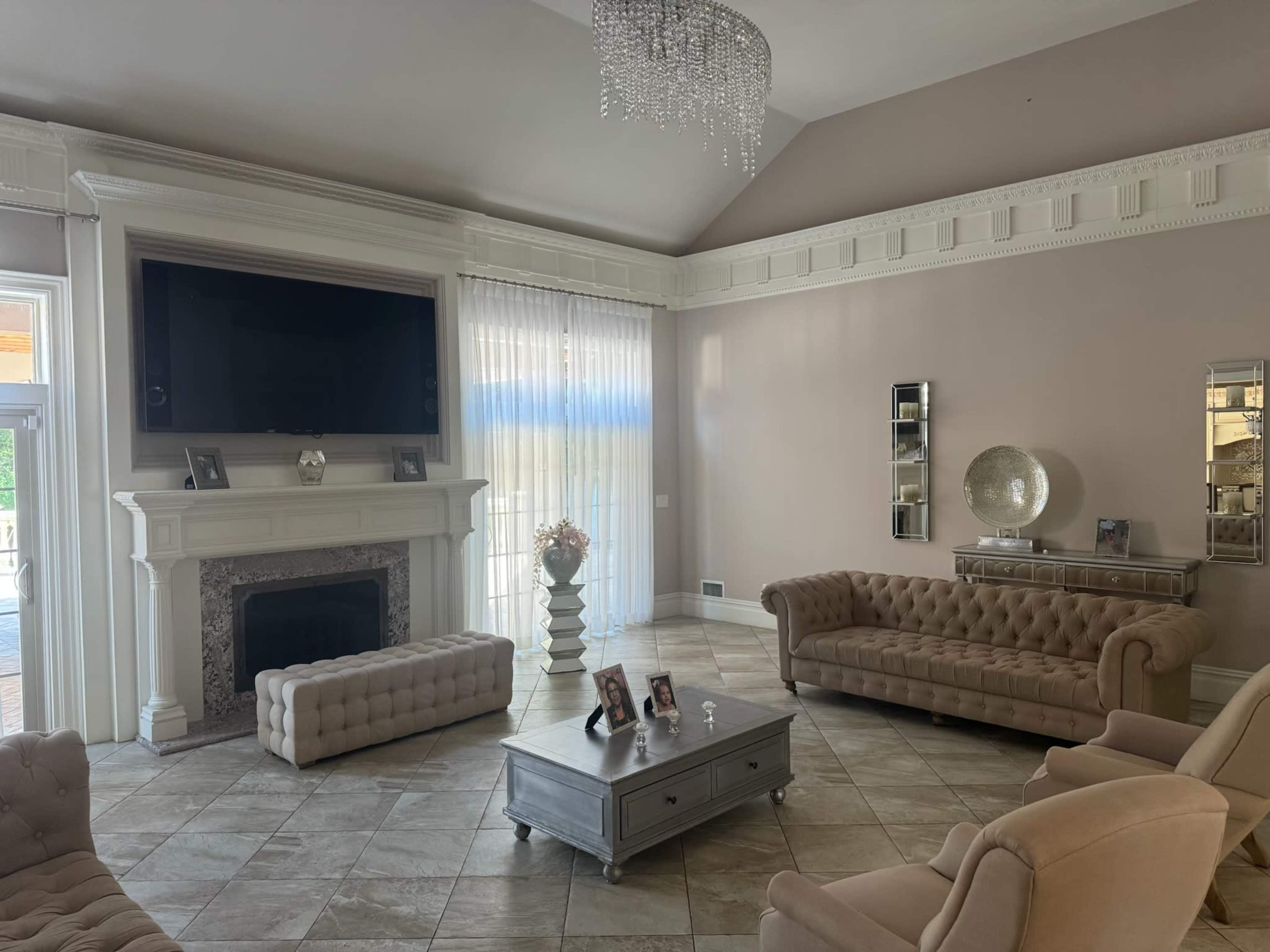 The living room features a high ceiling, a large flat-screen TV mounted above a fireplace, and two beige tufted sofas arranged around a coffee table on a tiled floor.