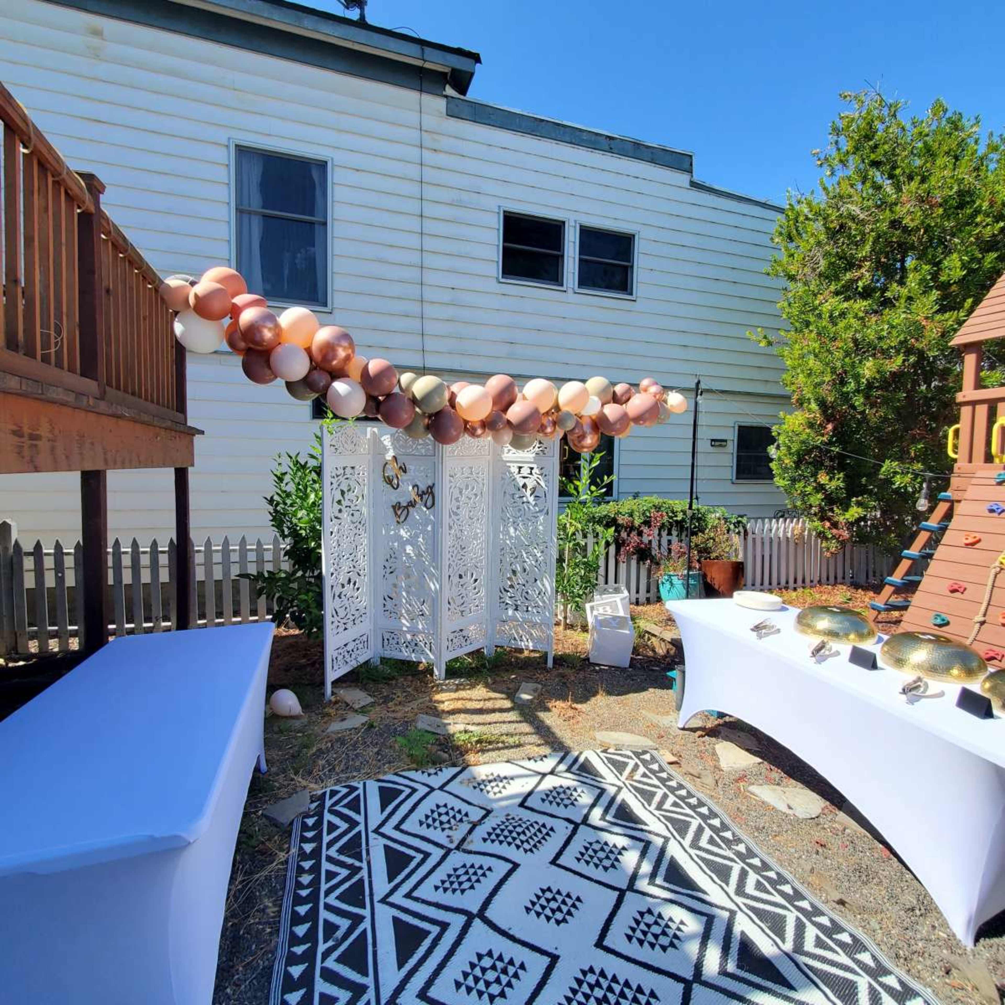 The image shows a decorated outdoor space featuring a balloon garland, a decorative screen, and two tables with items on display, set against a backdrop of a building and greenery.