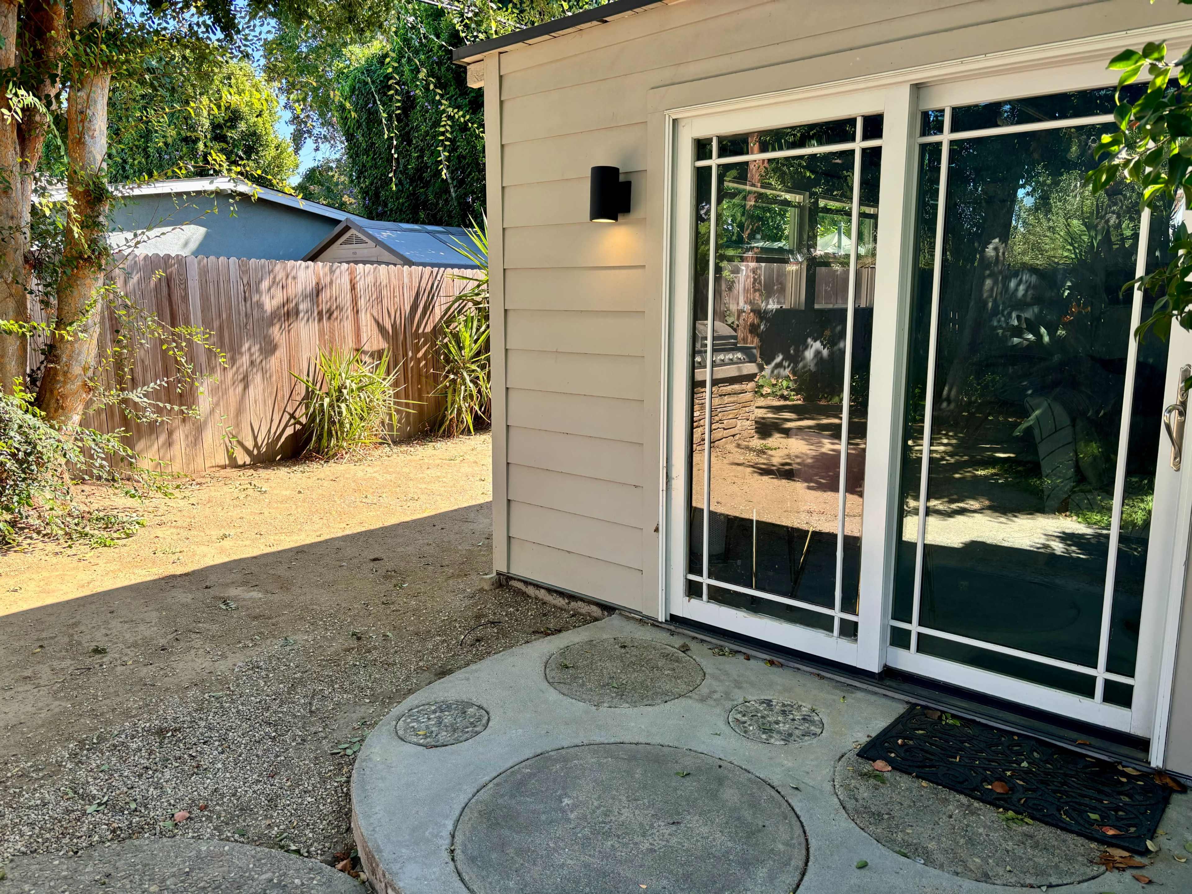A pathway leads to a glass door on a house, with a fenced yard and trees in the background.