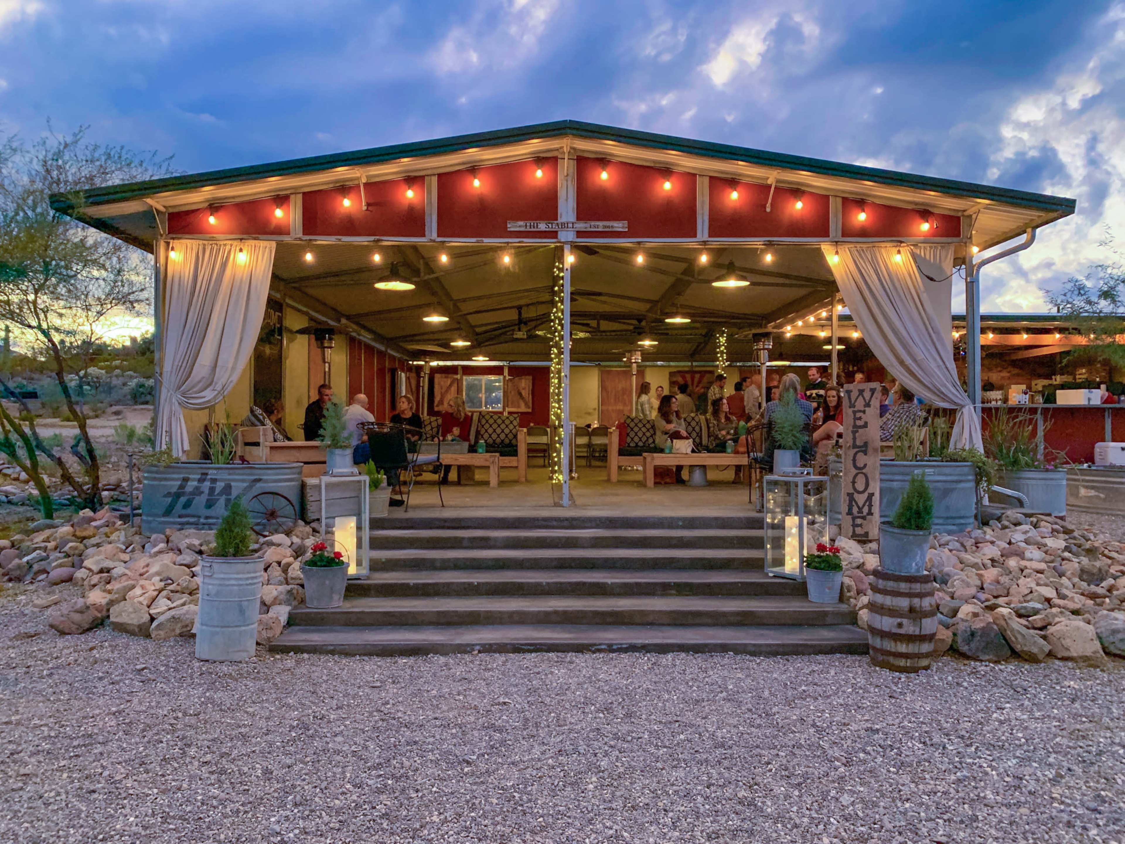 A rustic outdoor venue with a covered patio, adorned with string lights and hosting a gathering of people, is surrounded by desert scenery.
