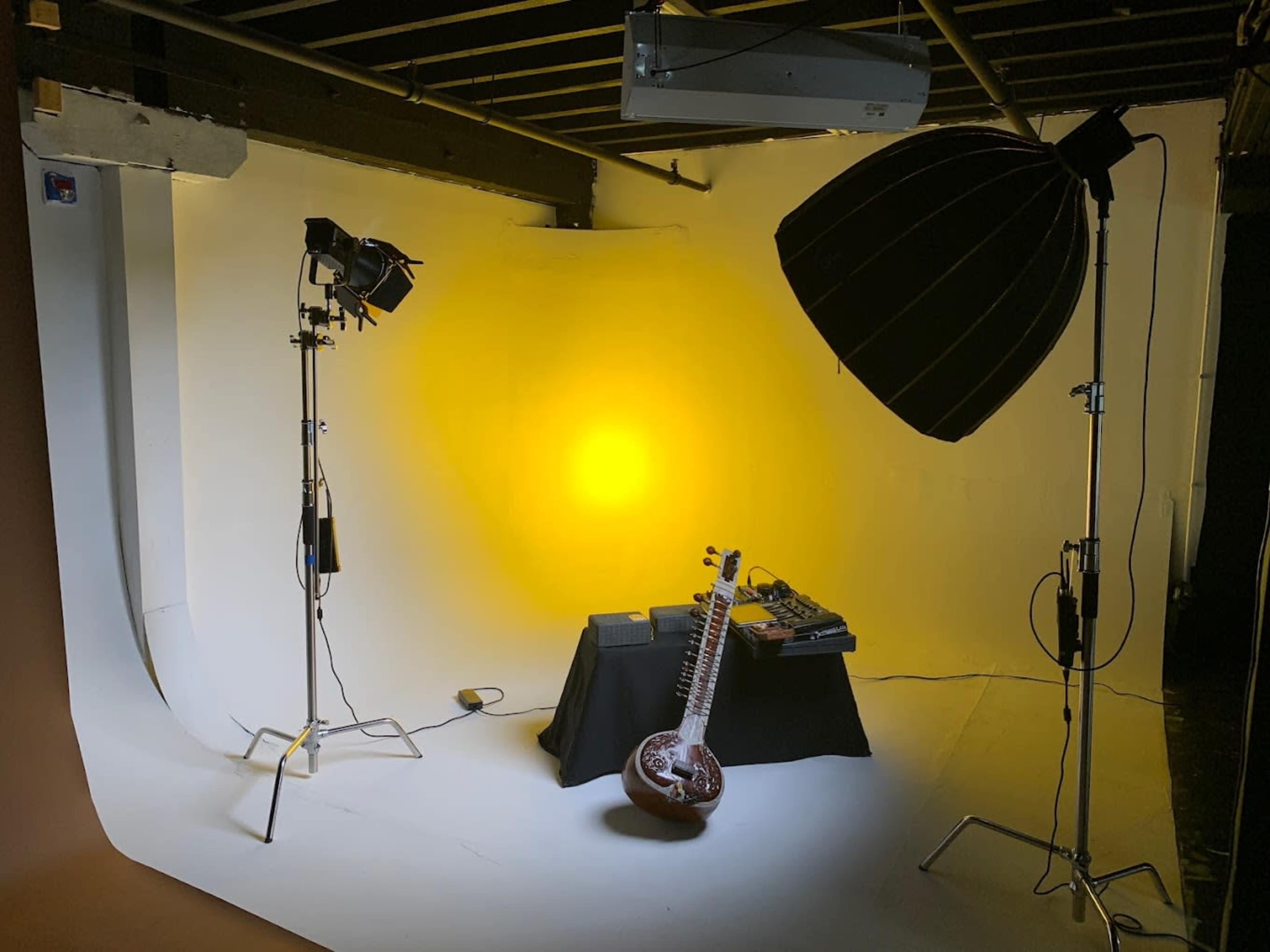 A photography studio setup featuring a sitar placed on a table, accompanied by music equipment, with two light sources illuminating a yellow backdrop.