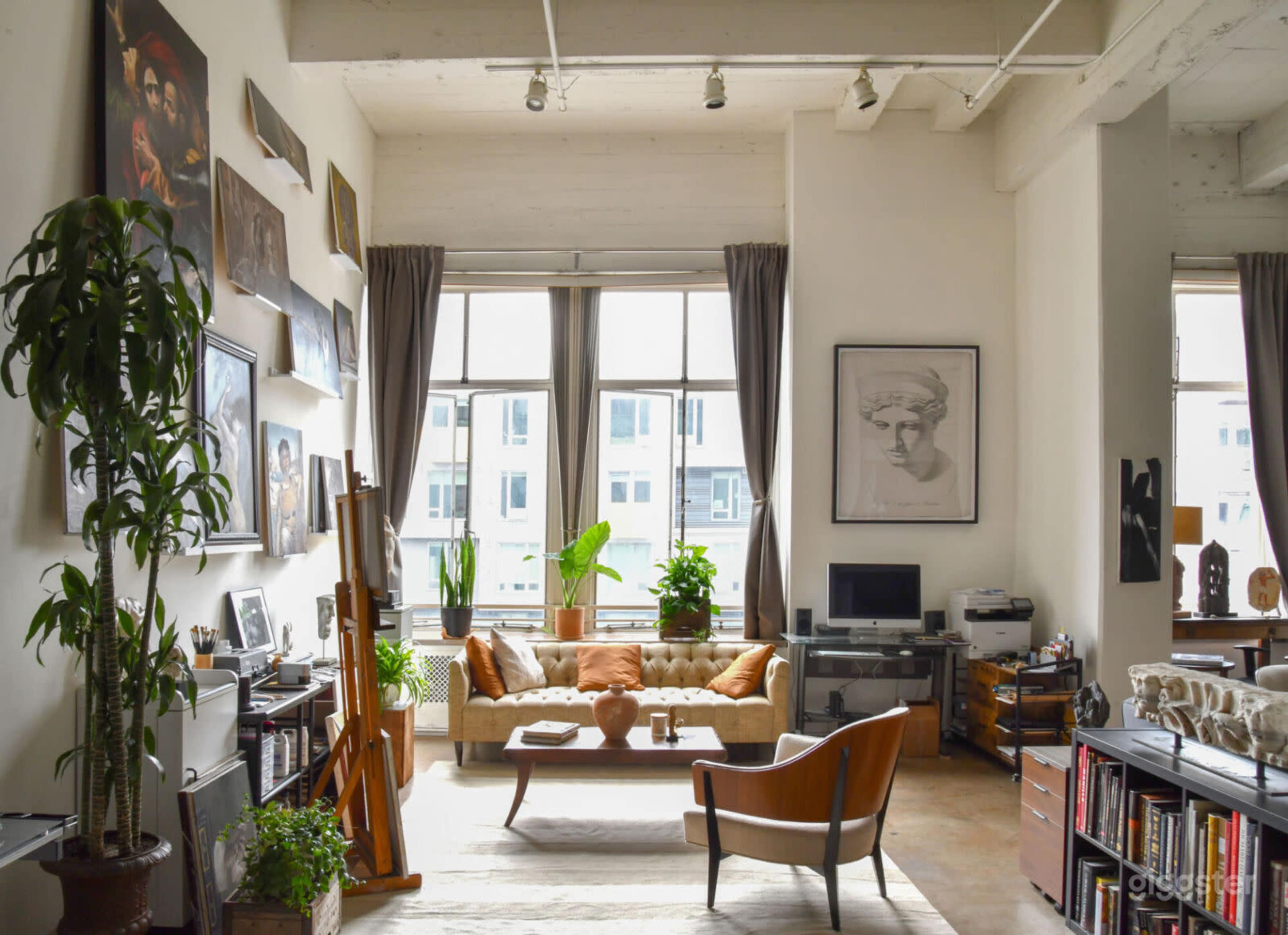 A spacious living room features large windows, a gallery wall of artwork, a couch with cushions, and various plants, creating an inviting atmosphere.